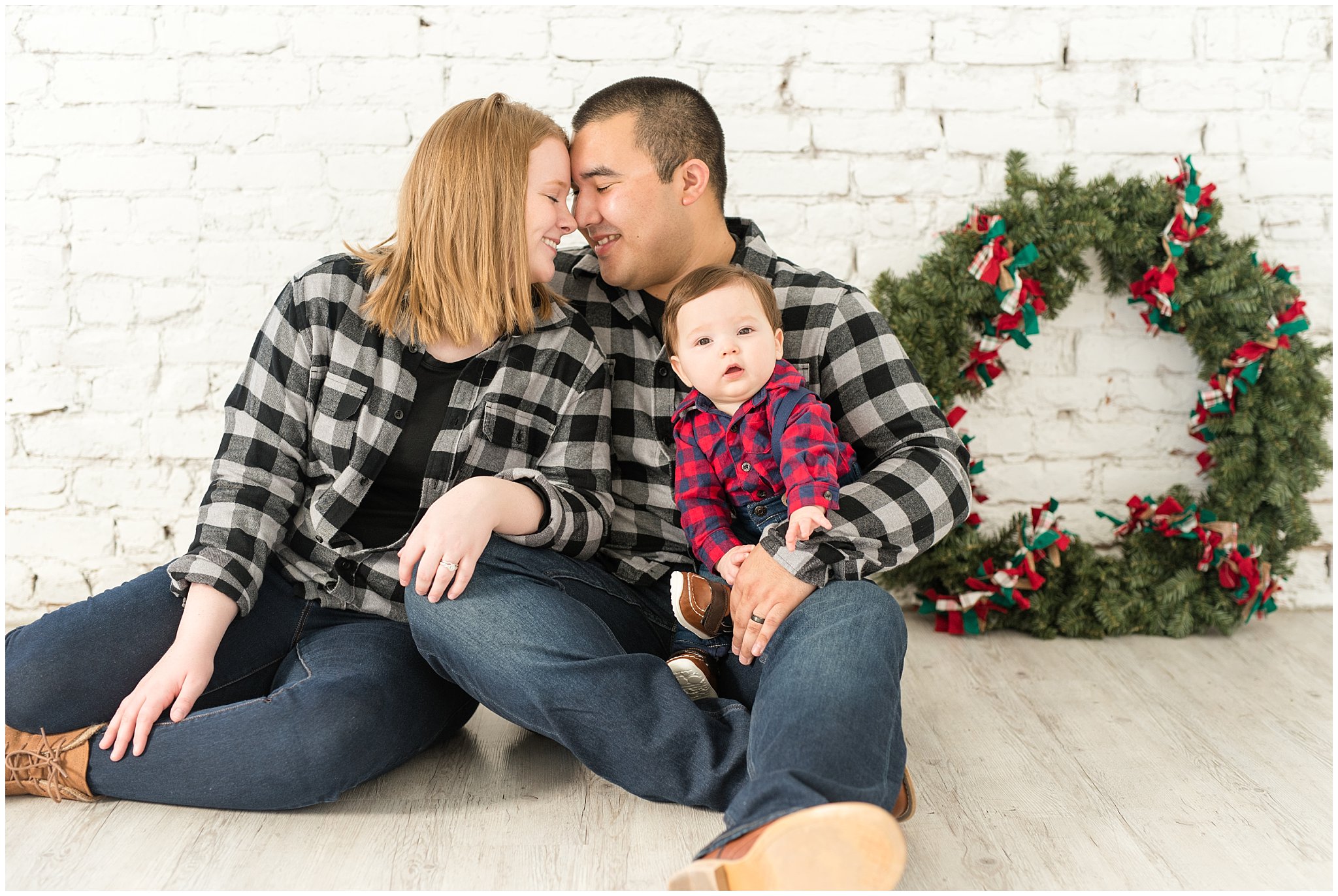 Parents nose to nose and holding baby boy in their arms with wreath behind them | Family Christmas session at the 5th floor | Utah Photographers | Jessie and Dallin Photography
