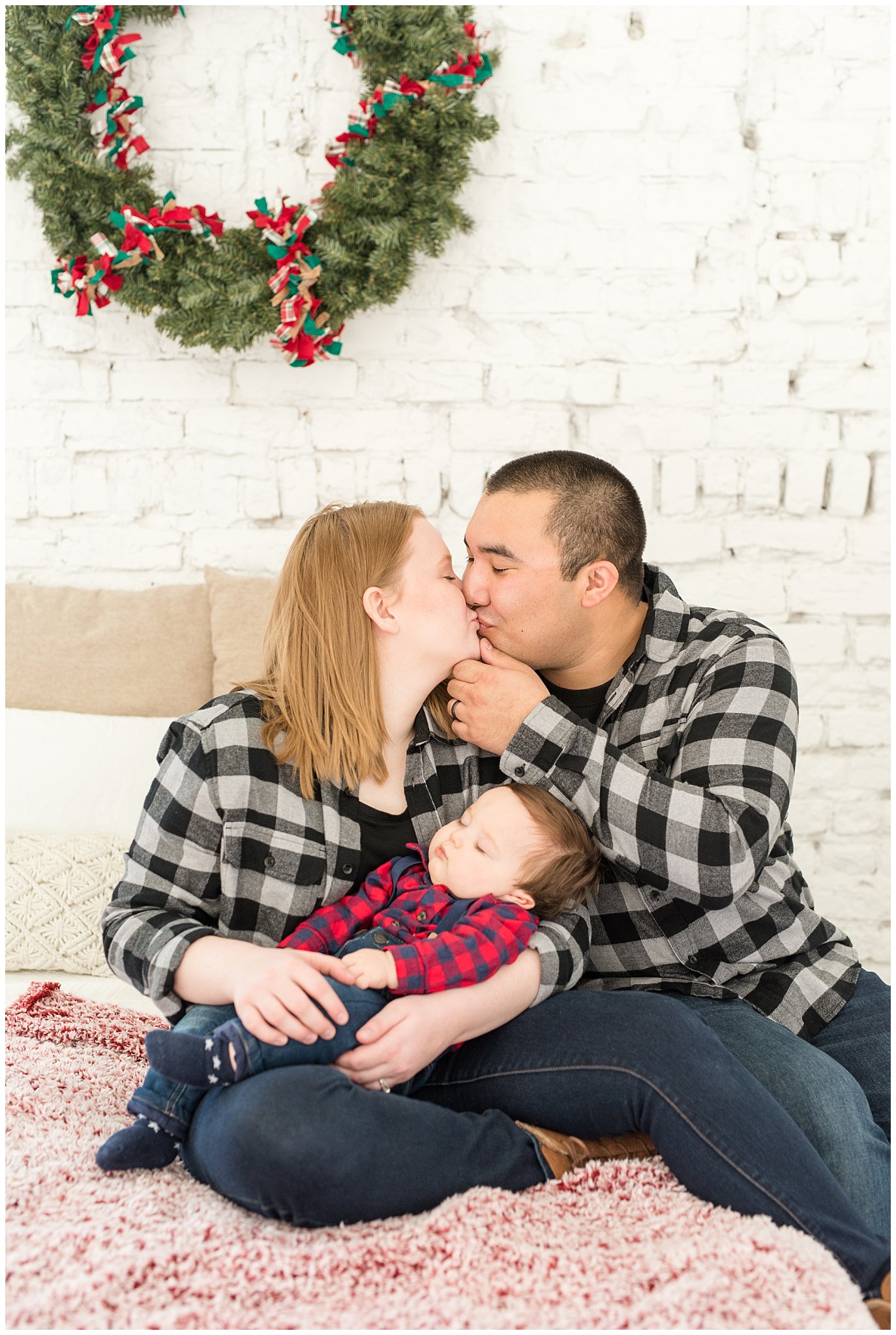 Parents kiss as baby boy sleeps in arms with wreath behind them on the bed | Family Christmas session at the 5th floor | Utah Photographers | Jessie and Dallin Photography