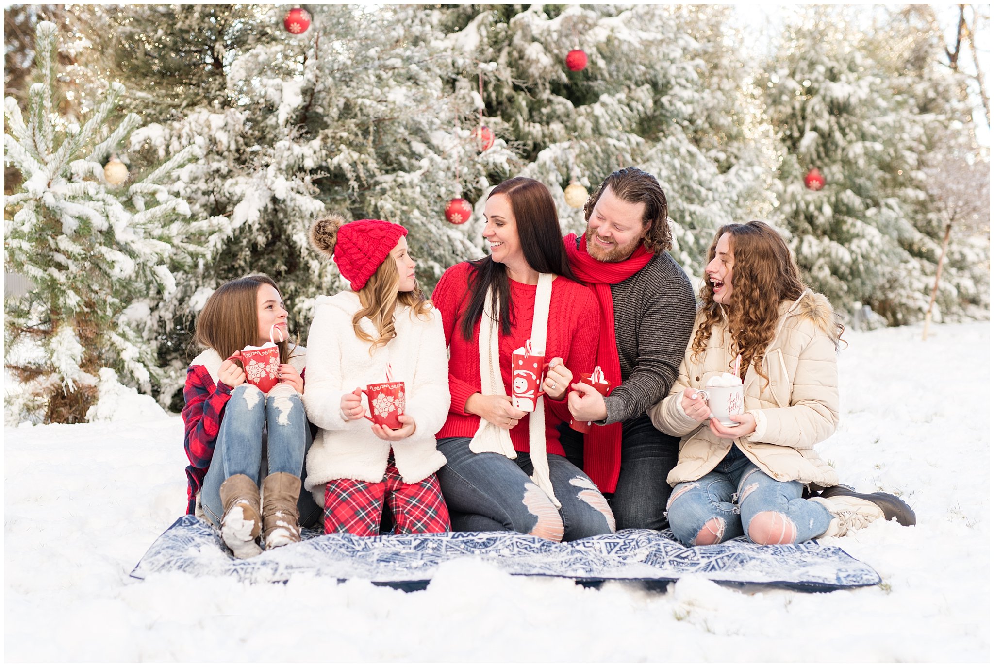 Family laughing in front of snowy Christmas trees holding hot chocolate mugs | Utah Family Christmas Photoshoot | Oak Hills Reception and Event | Jessie and Dallin Photography