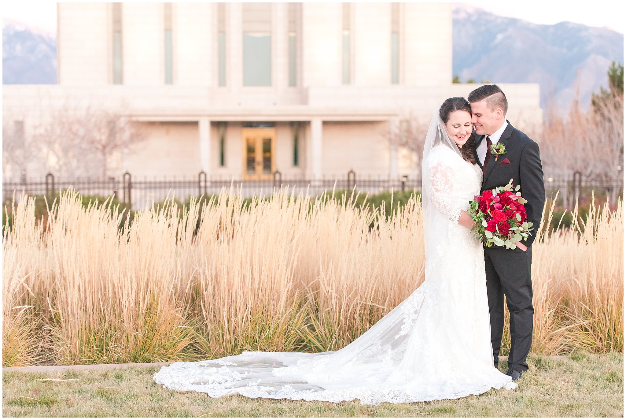 Bride in lace dress and veil with red floral Christmas theme bouquet and groom in black suit with burgundy tie | Utah mountains purple at sunset | oquirrh mountain temple winter formal session | Jessie and Dallin Photography