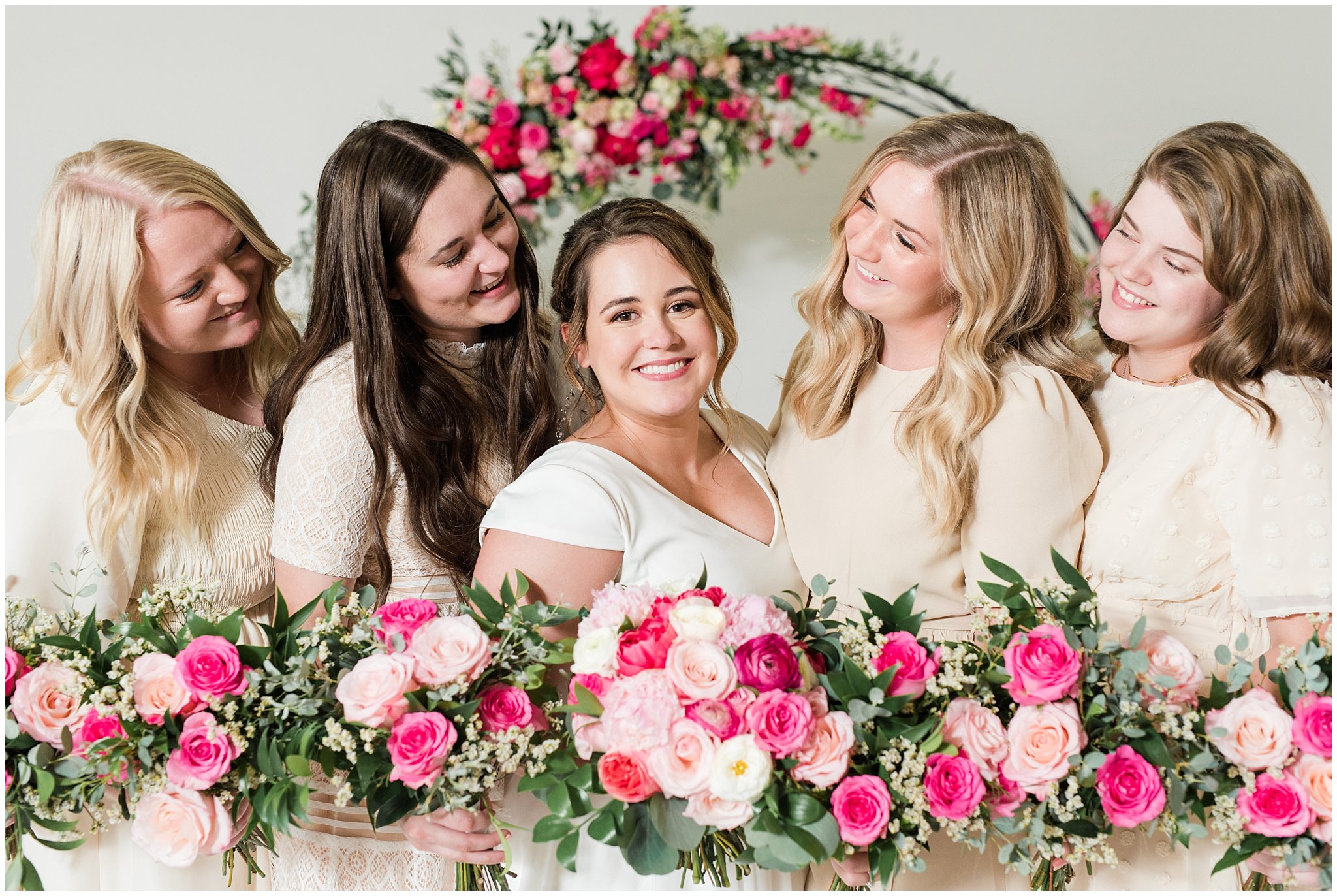 Bride and bridesmaids wearing cream colored dresses in front of floral arch in shades of pink | Talia Event Center Summer Wedding | Jessie and Dallin Photography