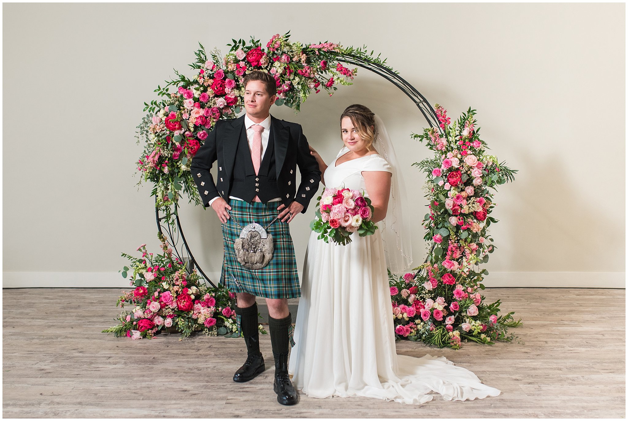 Bride and Groom in front of floral arch in shades of pink wearing flowy dress and highland attire with kilt | Talia Event Center Summer Wedding | Jessie and Dallin Photography
