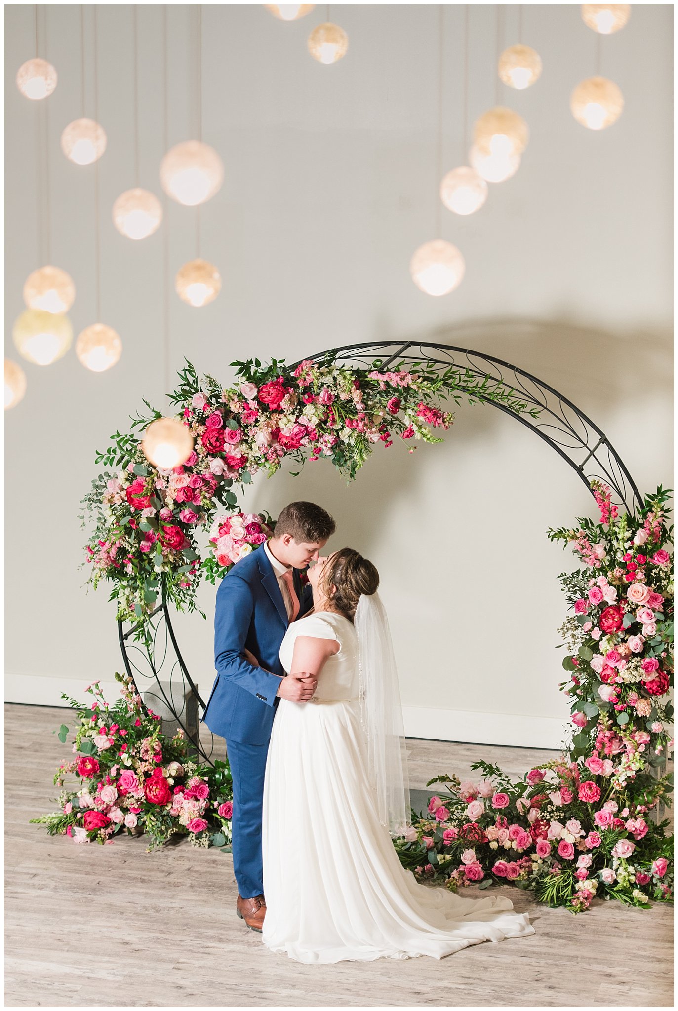 Bride and Groom in front of floral arch in shades of pink wearing flowy dress and cornish blue suit | Talia Event Center Summer Wedding | Jessie and Dallin Photography