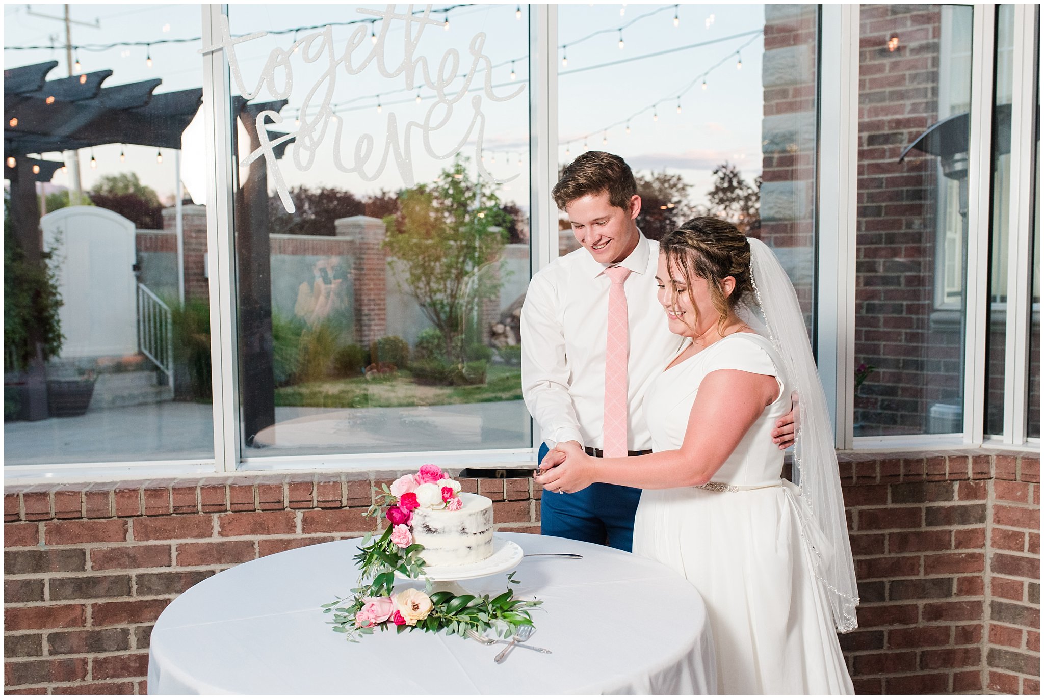 Bride and groom cut cookies and cream cake in front of windows and covered with pink and white florals | Talia Event Center Summer Wedding | Jessie and Dallin Photography