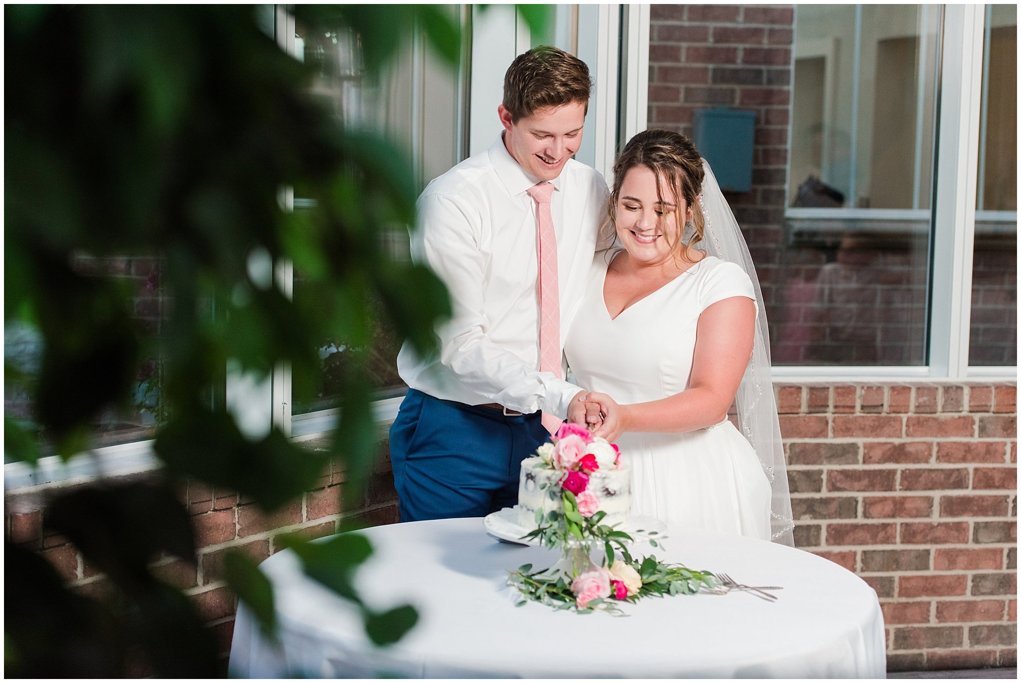 Bride and groom cut cookies and cream cake in front of windows and covered with pink and white florals | Talia Event Center Summer Wedding | Jessie and Dallin Photography