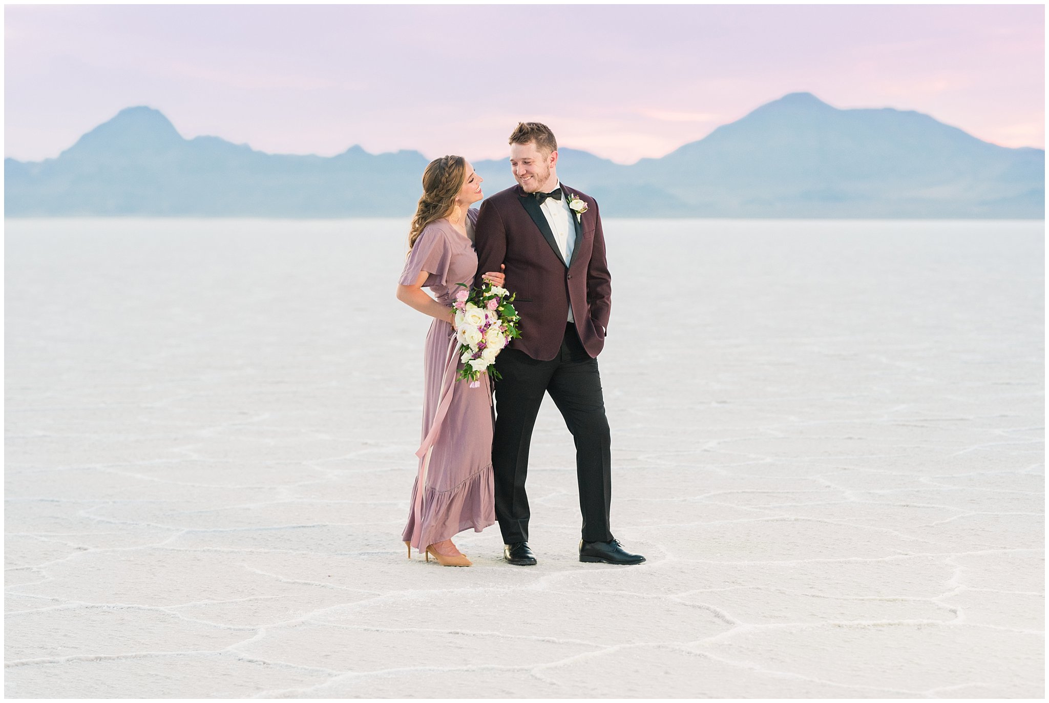 Couple during sunset wearing a dusty rose dress and burgundy suit with white and pink bouquet at the Bonneville Salt Flats | Bonneville Salt Flats Milky Way Anniversary Session | Jessie and Dallin Photography