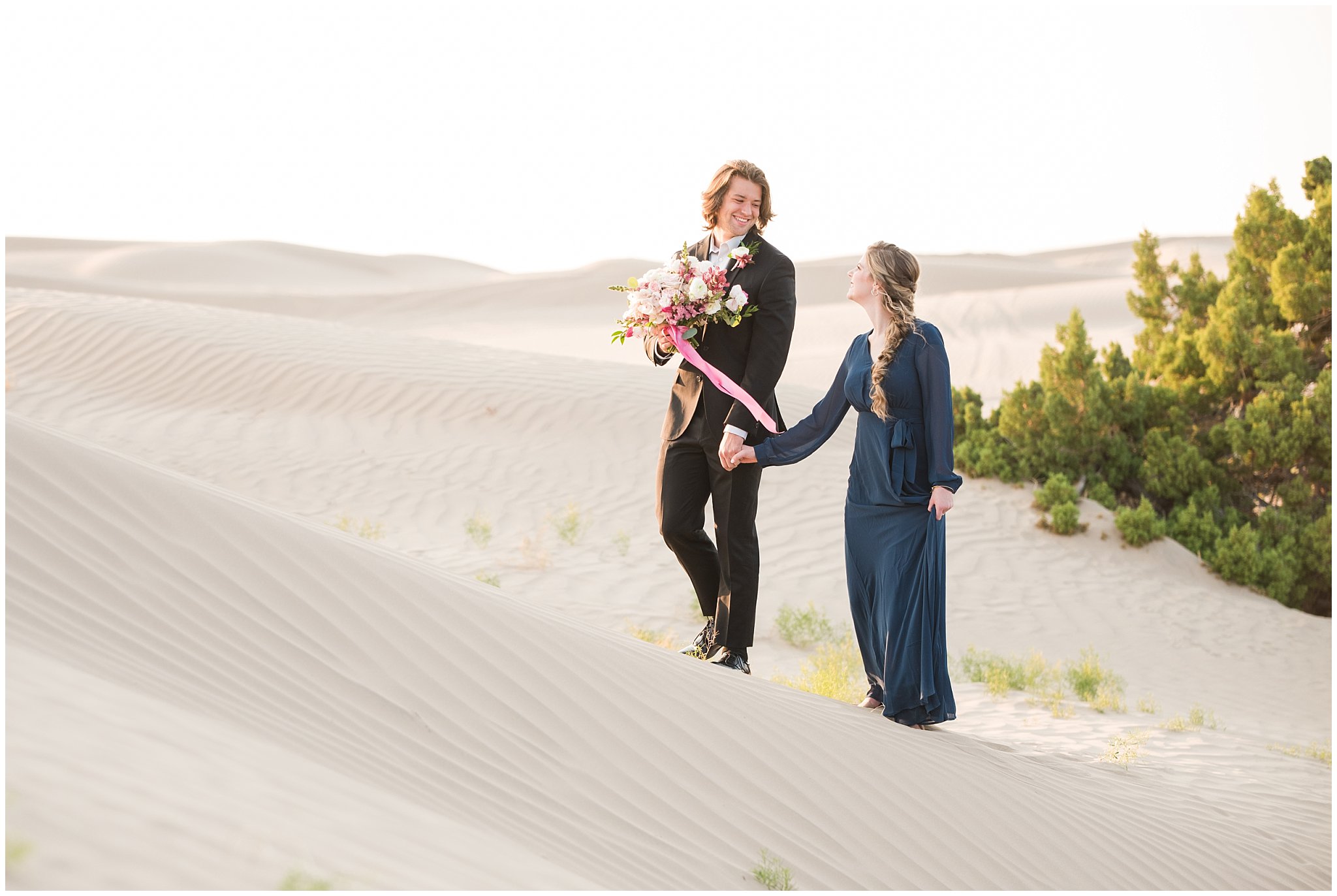 Couple dressed in black suit and blue flowy dress with white and pink orchid floral bouquet during couples session | Little Sahara Sand Dunes Milky Way Engagement Session