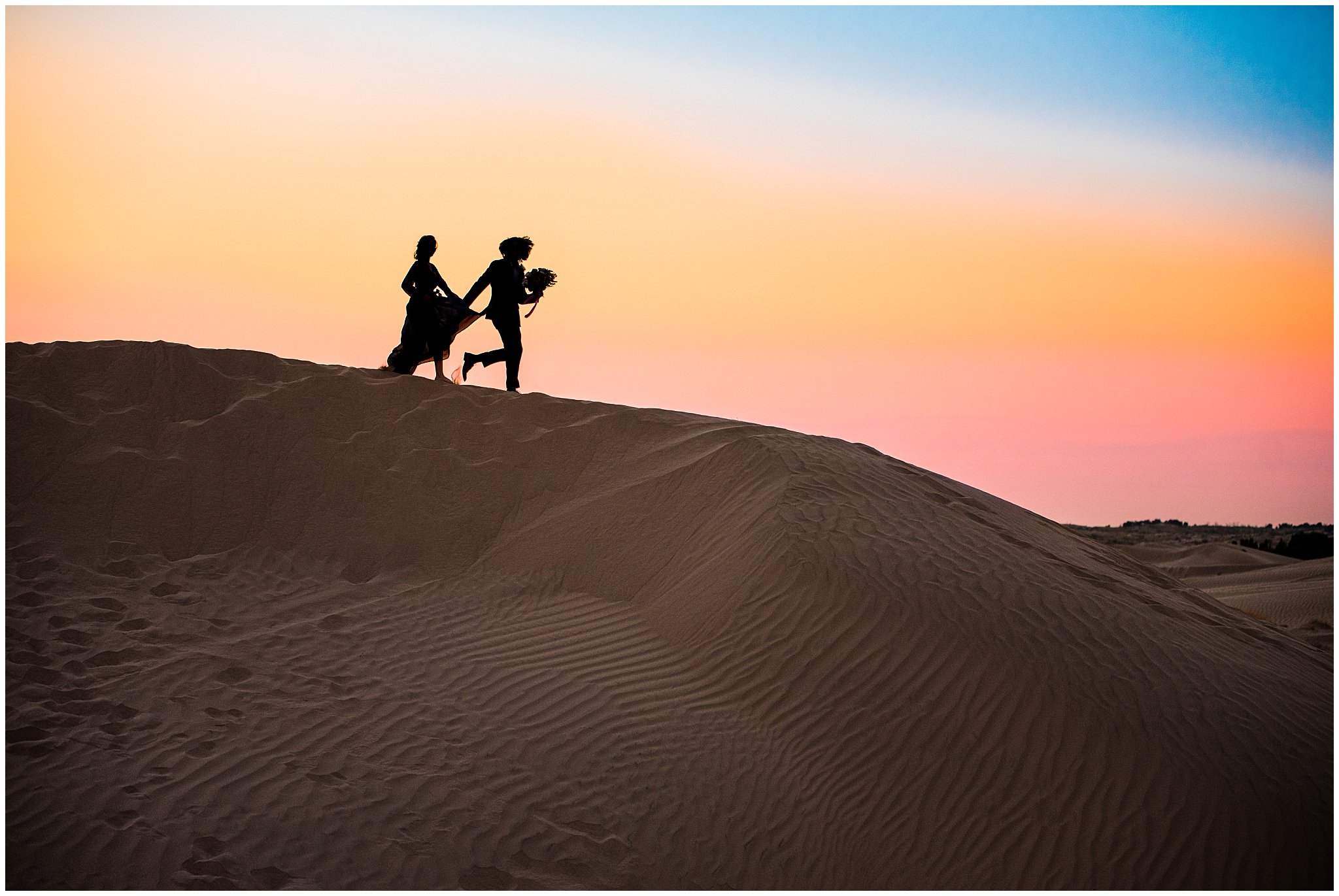 Couple at sunset dressed in black suit and blue flowy dress with white and pink orchid floral bouquet during couples session | Little Sahara Sand Dunes Milky Way Engagement Session
