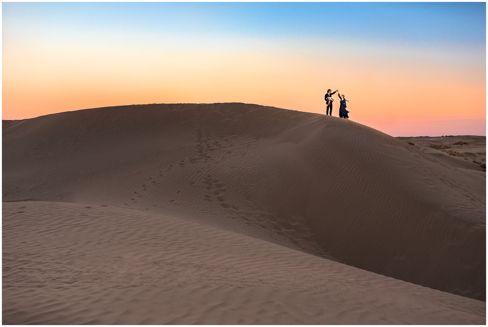 Couple at sunset dressed in black suit and blue flowy dress with white and pink orchid floral bouquet during couples session | Little Sahara Sand Dunes Milky Way Engagement Session