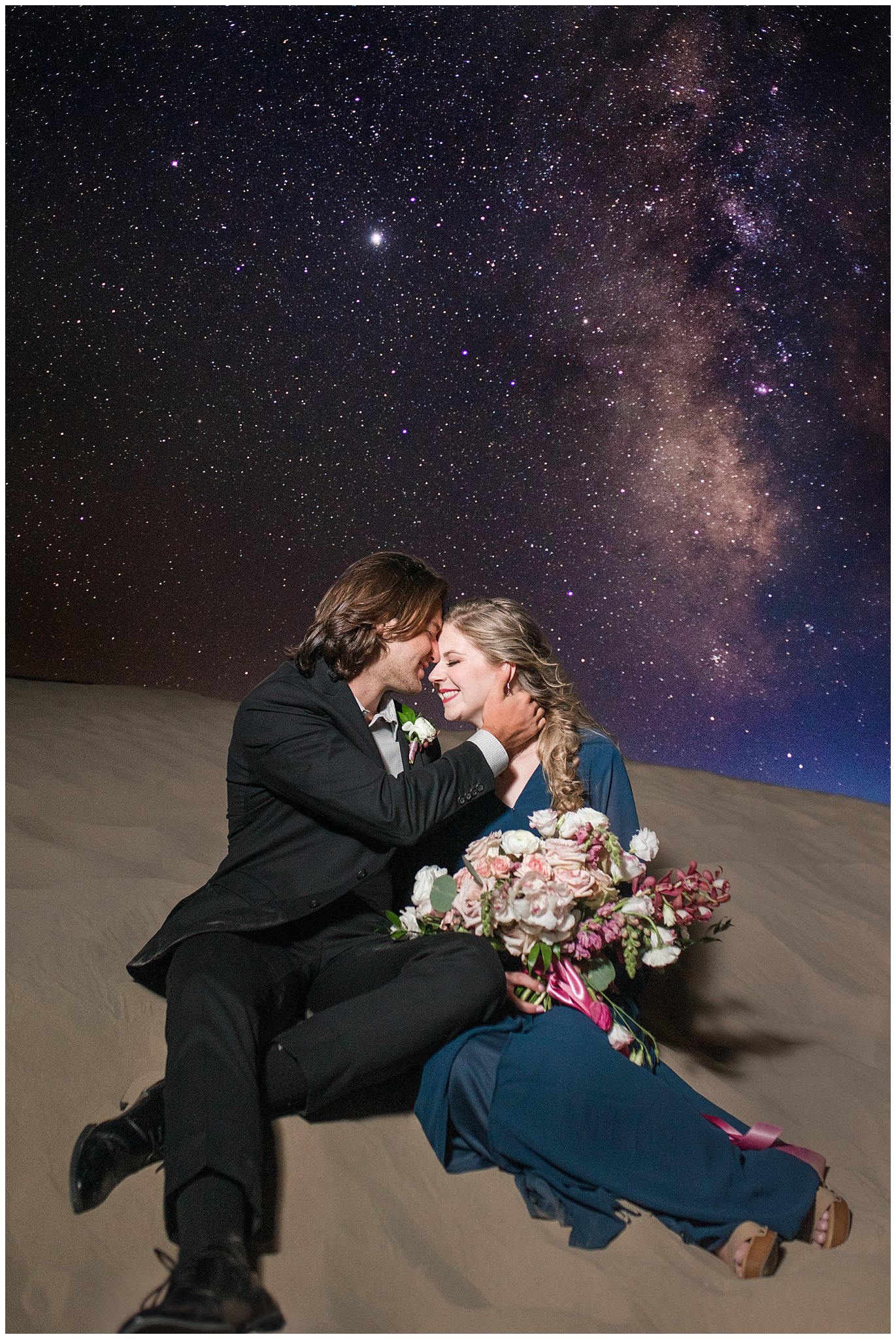 Couple with Milky Way galaxy dressed in black suit and blue flowy dress with white and pink orchid floral bouquet during couples session | Little Sahara Sand Dunes Milky Way Engagement Session