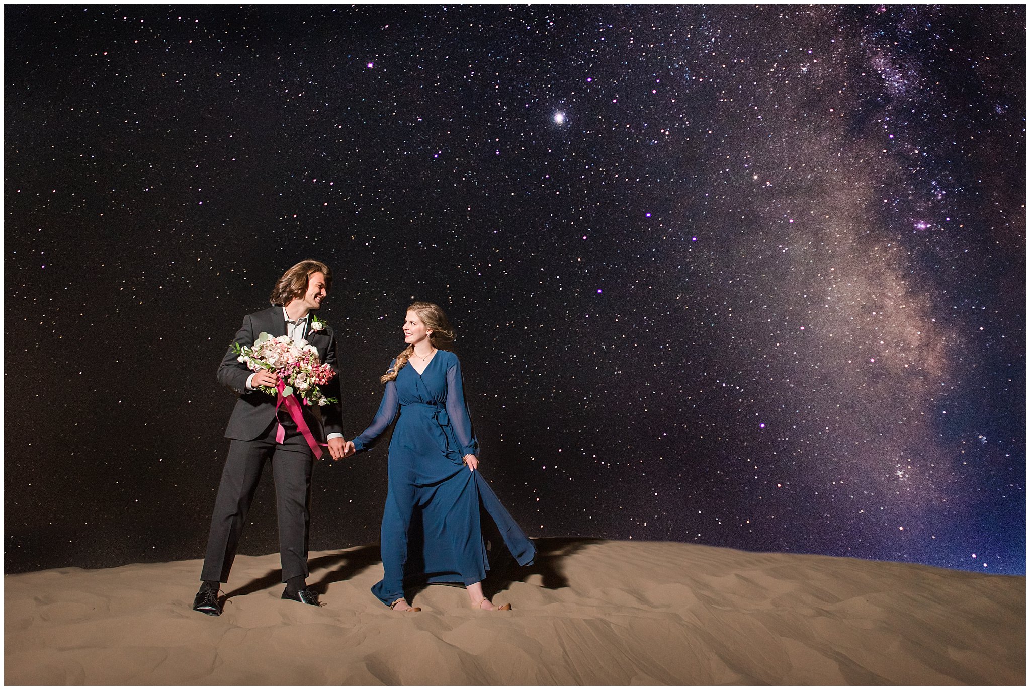 Couple with Milky Way galaxy dressed in black suit and blue flowy dress with white and pink orchid floral bouquet during couples session | Little Sahara Sand Dunes Milky Way Engagement Session