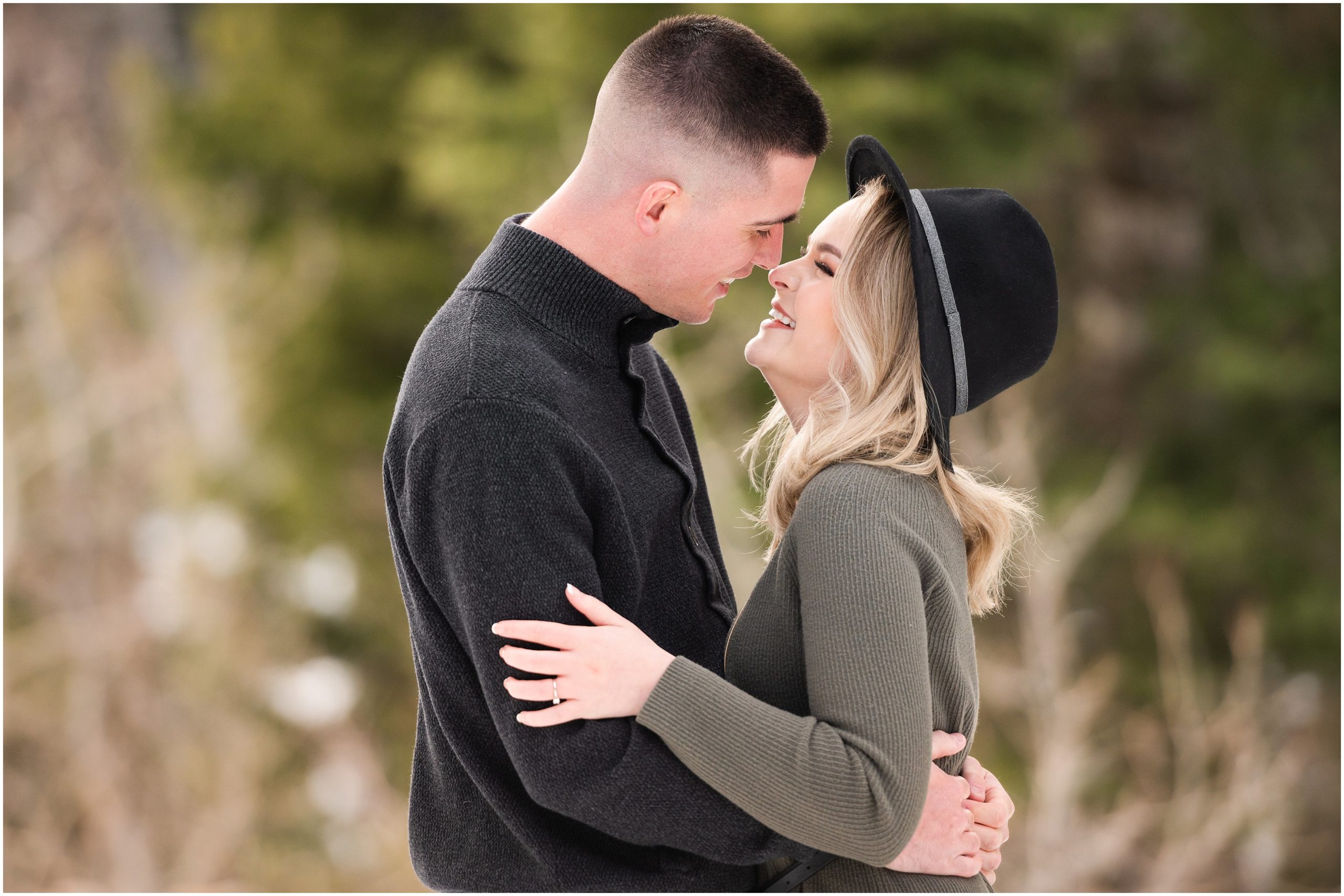 Couple in the snow during mountain destination engagement session in Utah | Wearing olive colored dress with black floppy hat and black sweater and black pants | Snowbasin Resort Snowy Engagement Session | Jessie and Dallin Photography