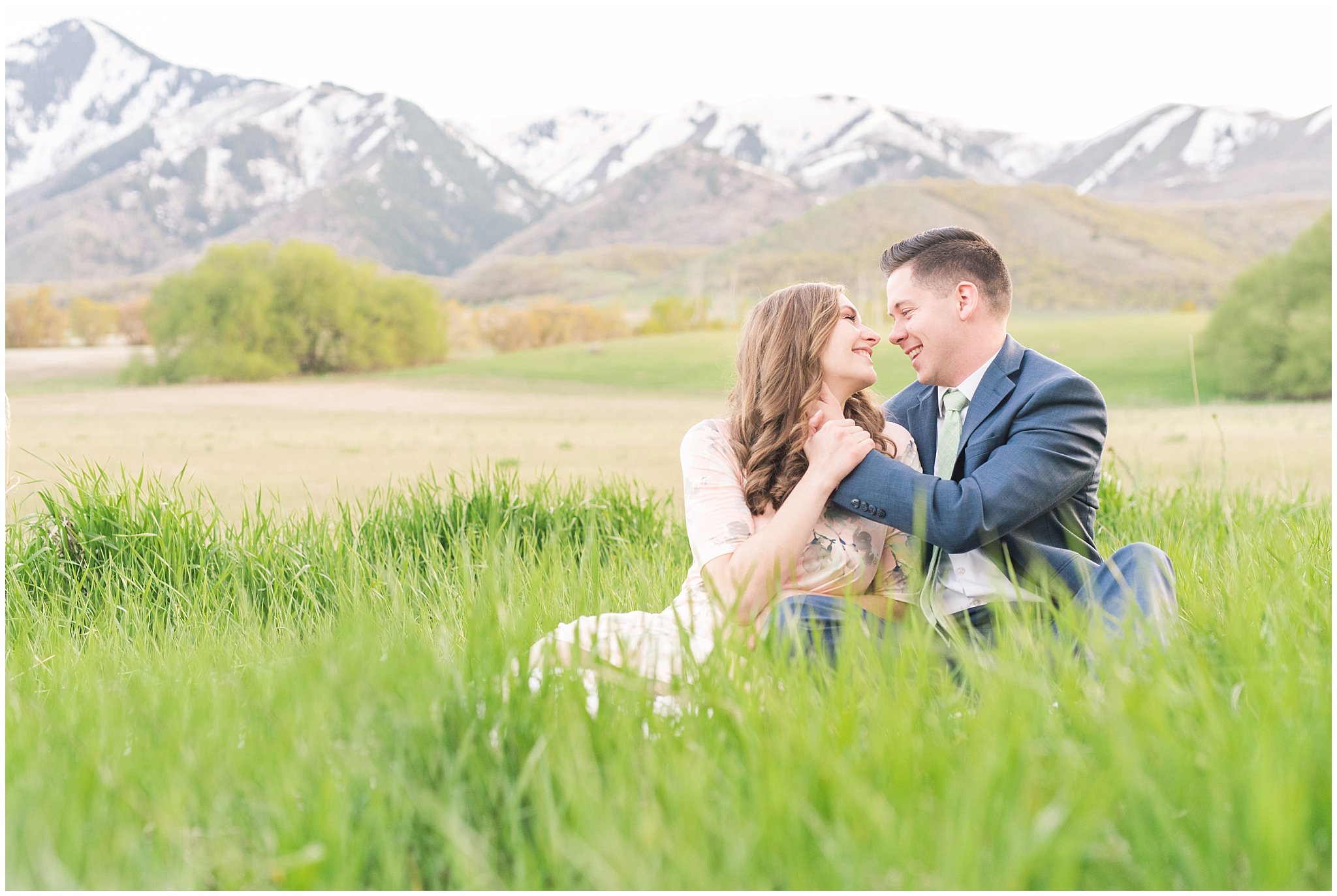 Couple dressed up in blue suit and blush dress surrounded by grass and snowy mountain peaks during elegant engagement | Downtown Logan and Wellsville Mountain Engagement | Jessie and Dallin Photography