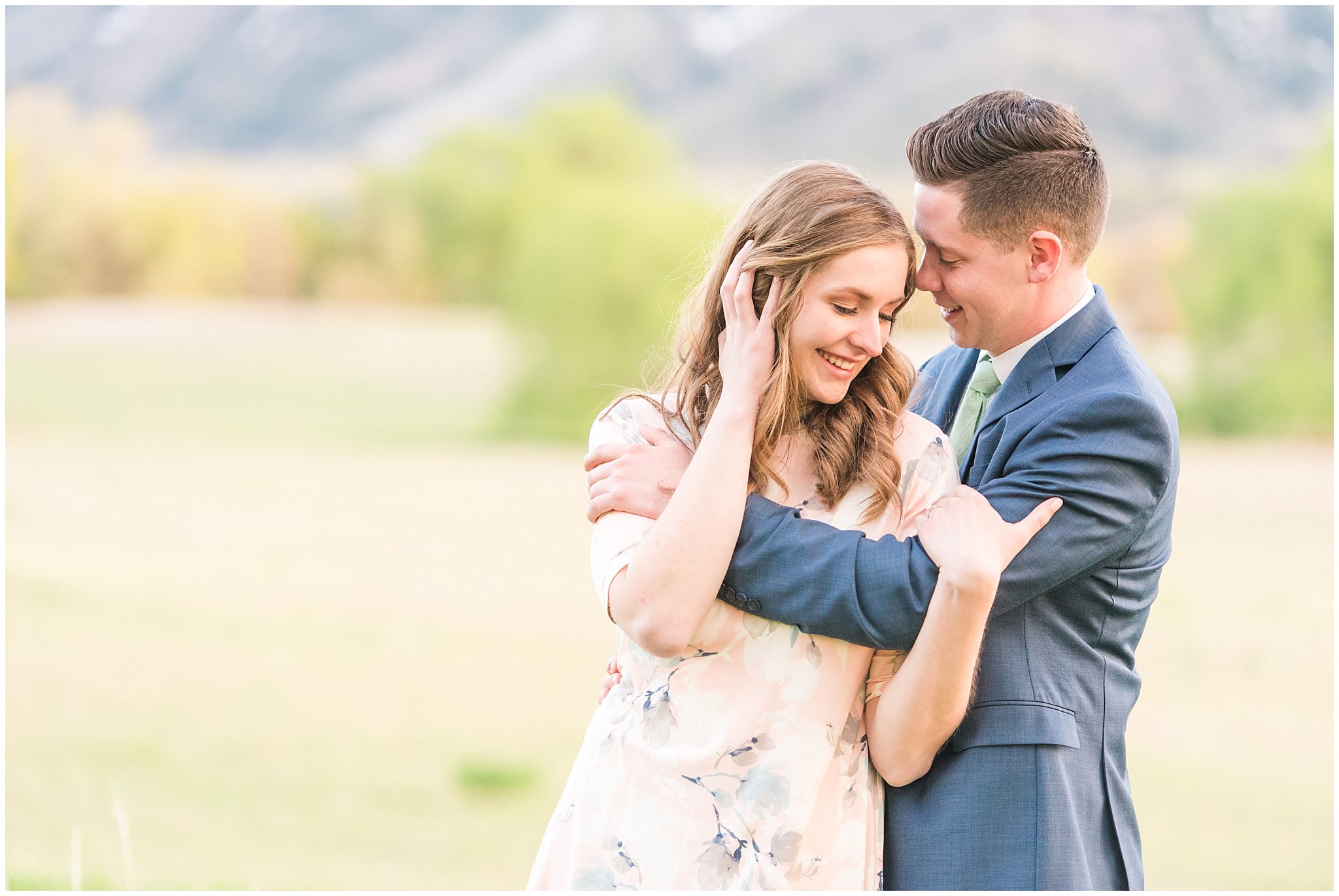 Couple dressed up in blue suit and blush dress surrounded by grass and snowy mountain peaks during elegant engagement | Downtown Logan and Wellsville Mountain Engagement | Jessie and Dallin Photography