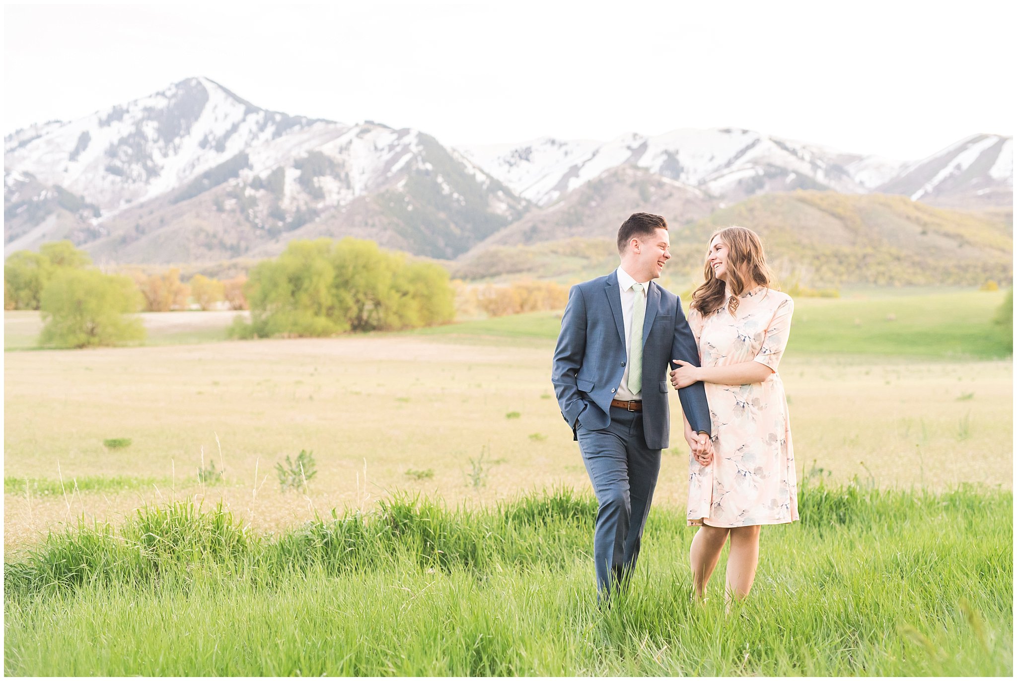 Couple dressed up in blue suit and blush dress surrounded by grass and snowy mountain peaks during elegant engagement | Downtown Logan and Wellsville Mountain Engagement | Jessie and Dallin Photography