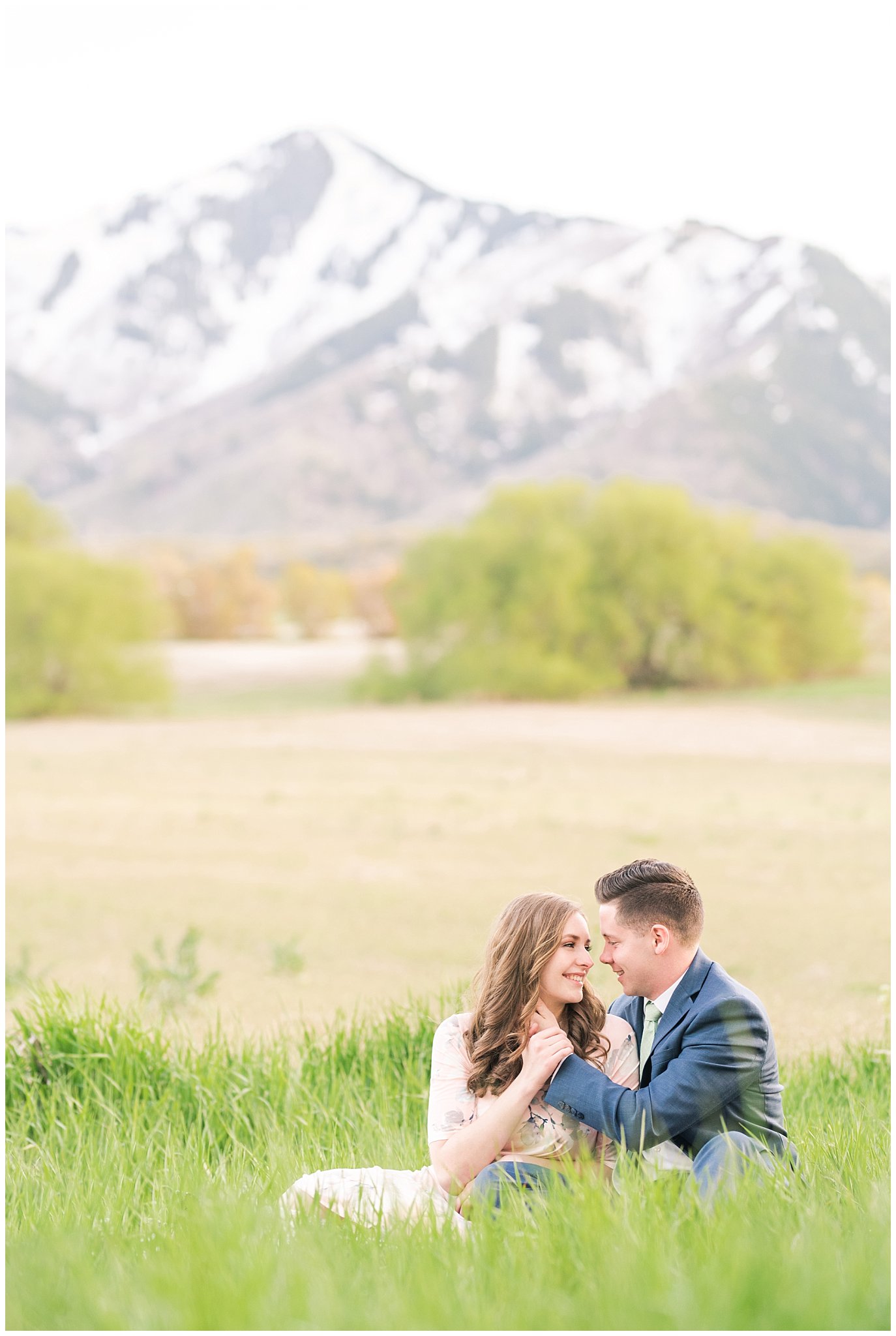 Couple dressed up in blue suit and blush dress surrounded by grass and snowy mountain peaks during elegant engagement | Downtown Logan and Wellsville Mountain Engagement | Jessie and Dallin Photography