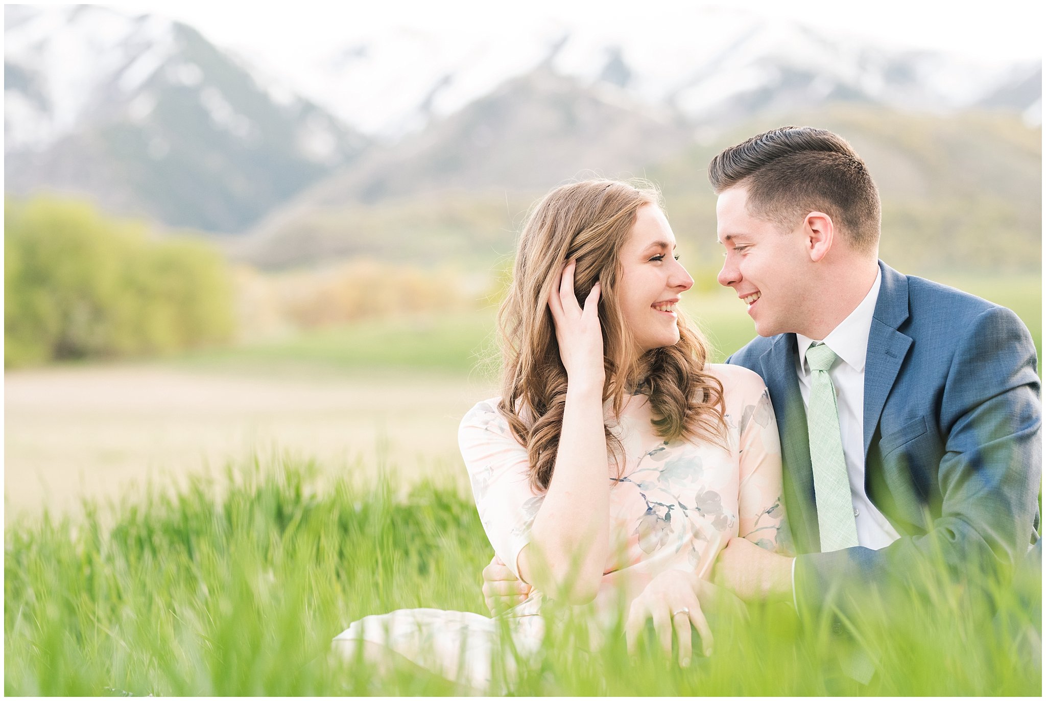 Couple dressed up in blue suit and blush dress surrounded by grass and snowy mountain peaks during elegant engagement | Downtown Logan and Wellsville Mountain Engagement | Jessie and Dallin Photography