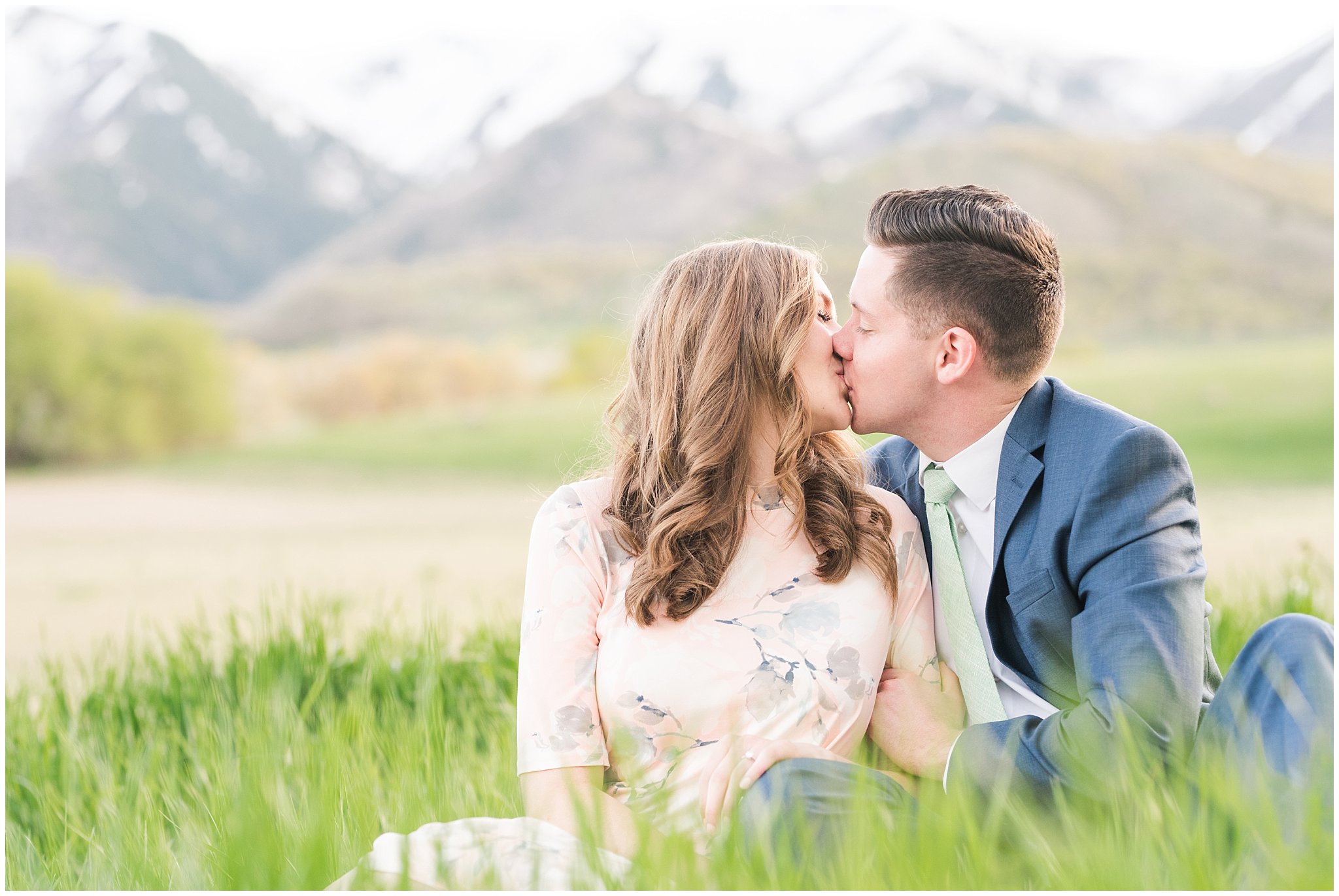 Couple dressed up in blue suit and blush dress surrounded by grass and snowy mountain peaks during elegant engagement | Downtown Logan and Wellsville Mountain Engagement | Jessie and Dallin Photography