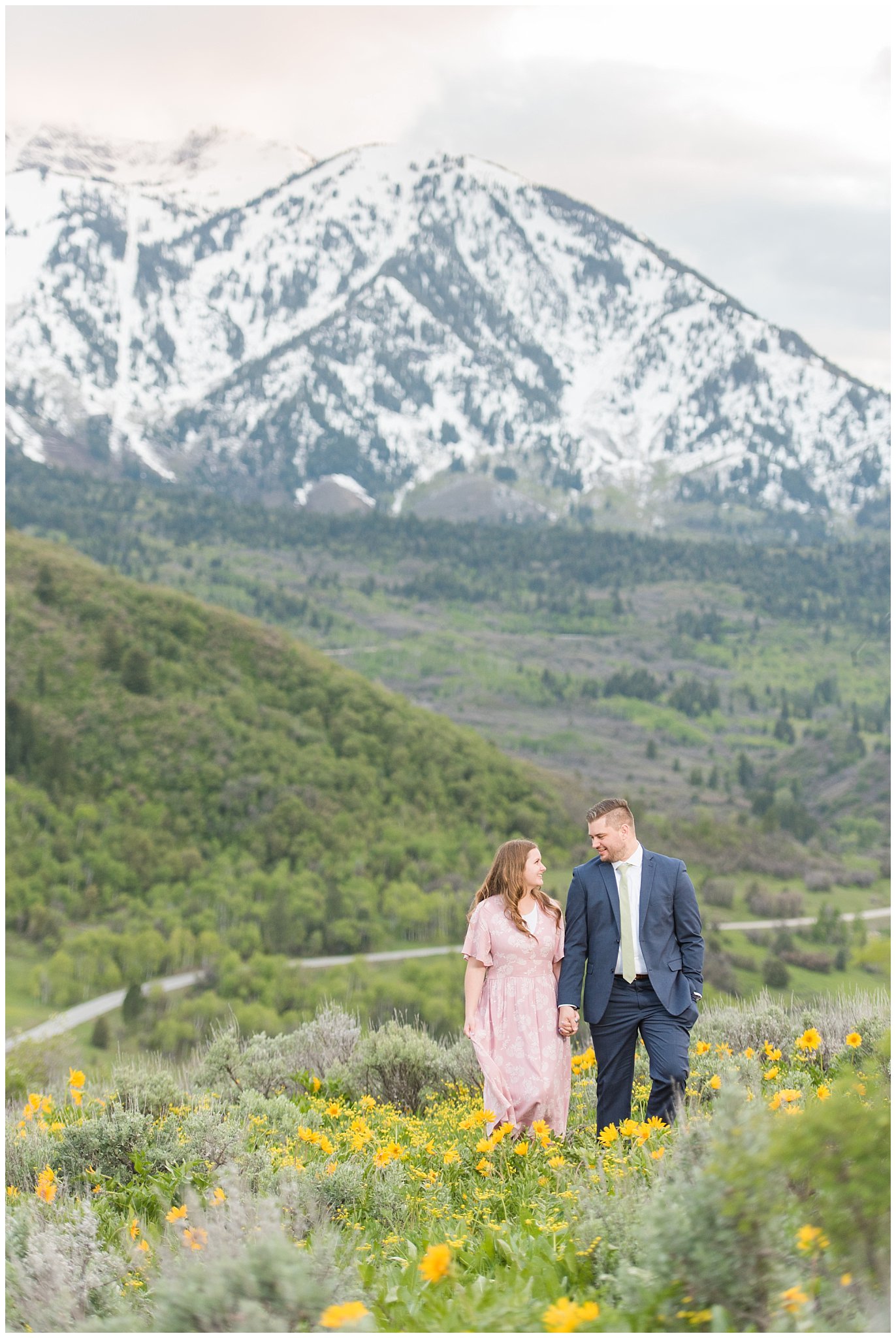 Couple dressed in blush dress and blue suit surrounded by sunflowers and snowy mountains | Ogden Valley Spring Mountain Engagement | Jessie and Dallin Photography