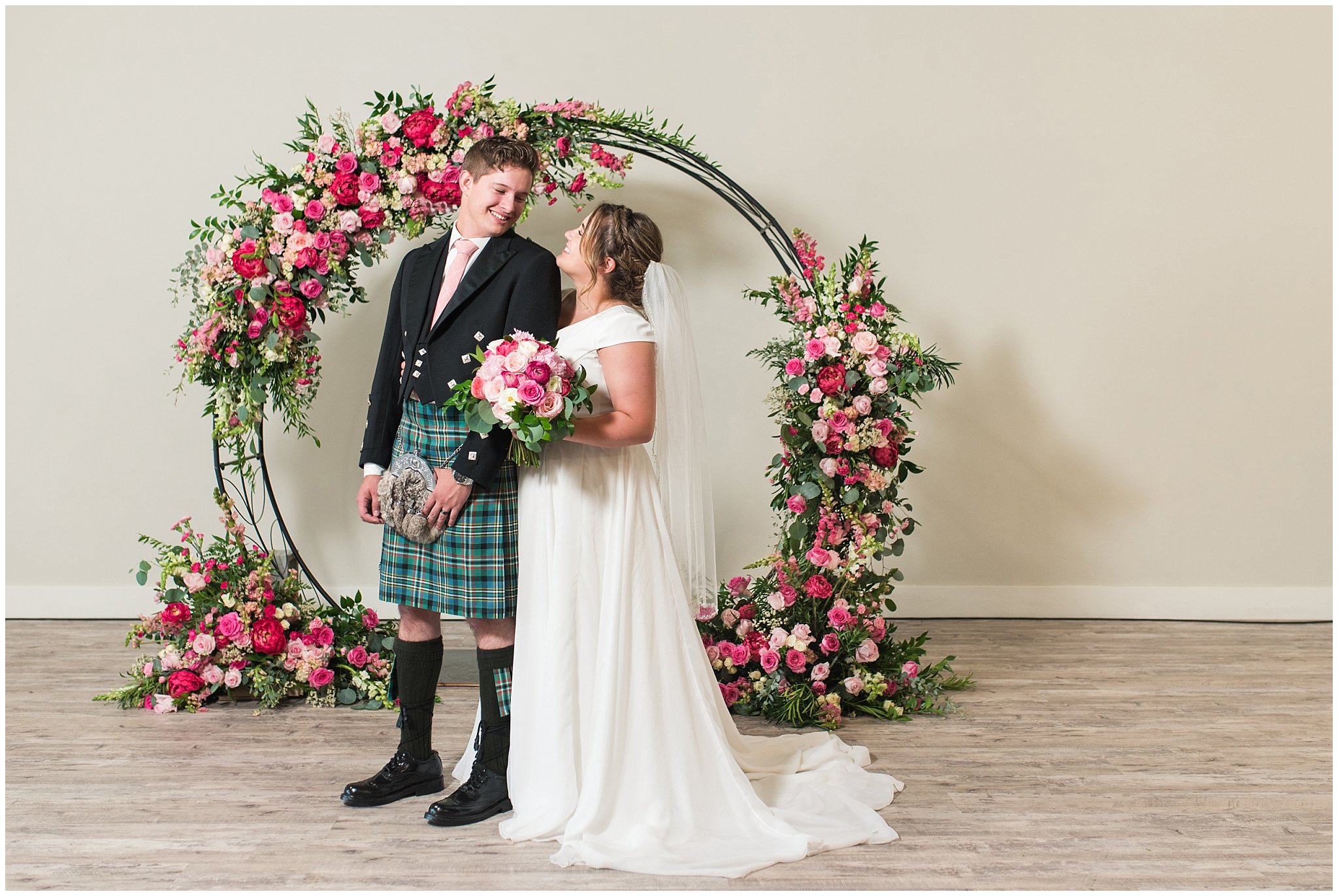 Bride and Groom in front of floral arch in shades of pink wearing flowy dress and highland attire with kilt | Talia Event Center Summer Wedding | Jessie and Dallin Photography