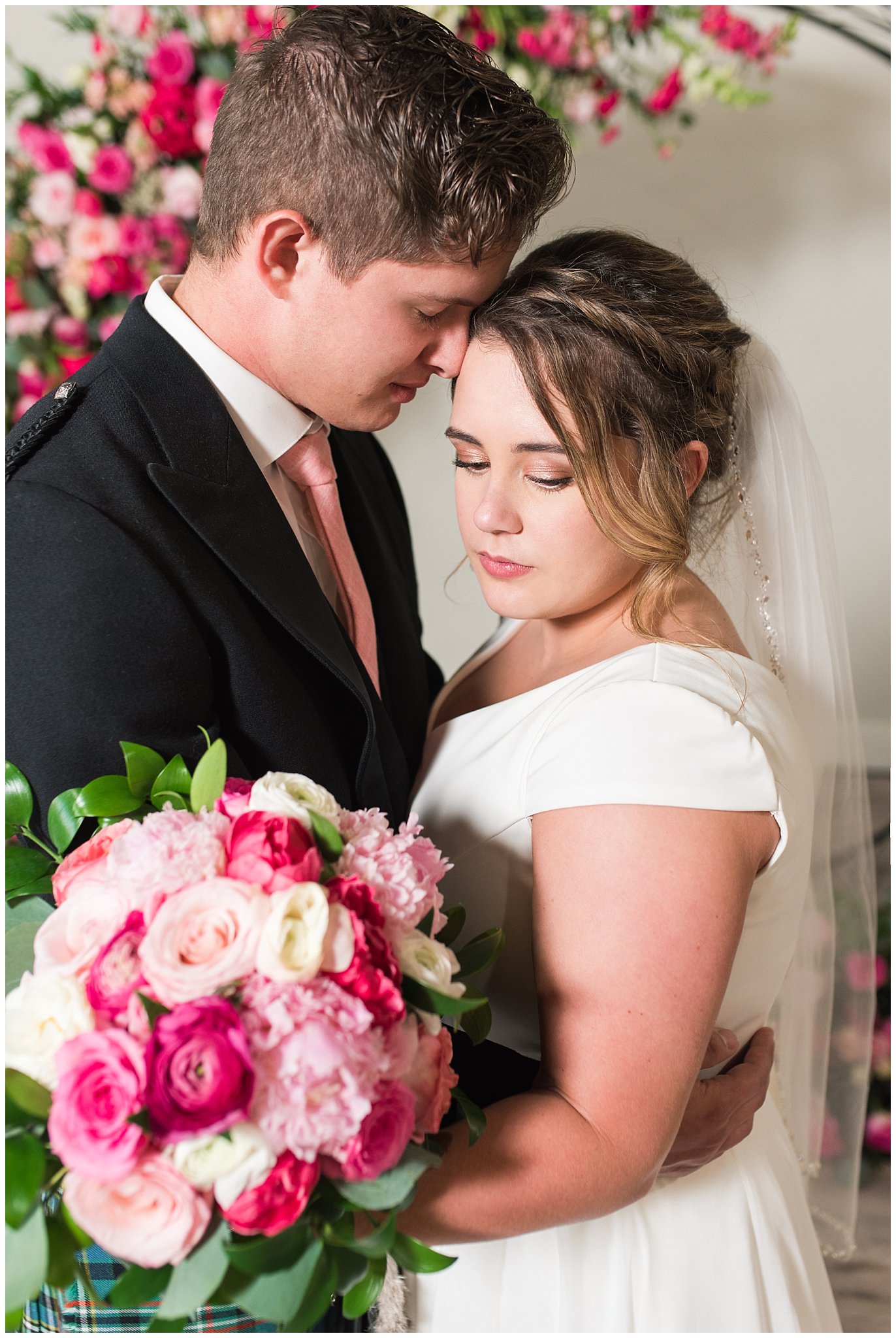 Bride and Groom in front of floral arch in shades of pink wearing flowy dress and highland attire with kilt | Talia Event Center Summer Wedding | Jessie and Dallin Photography