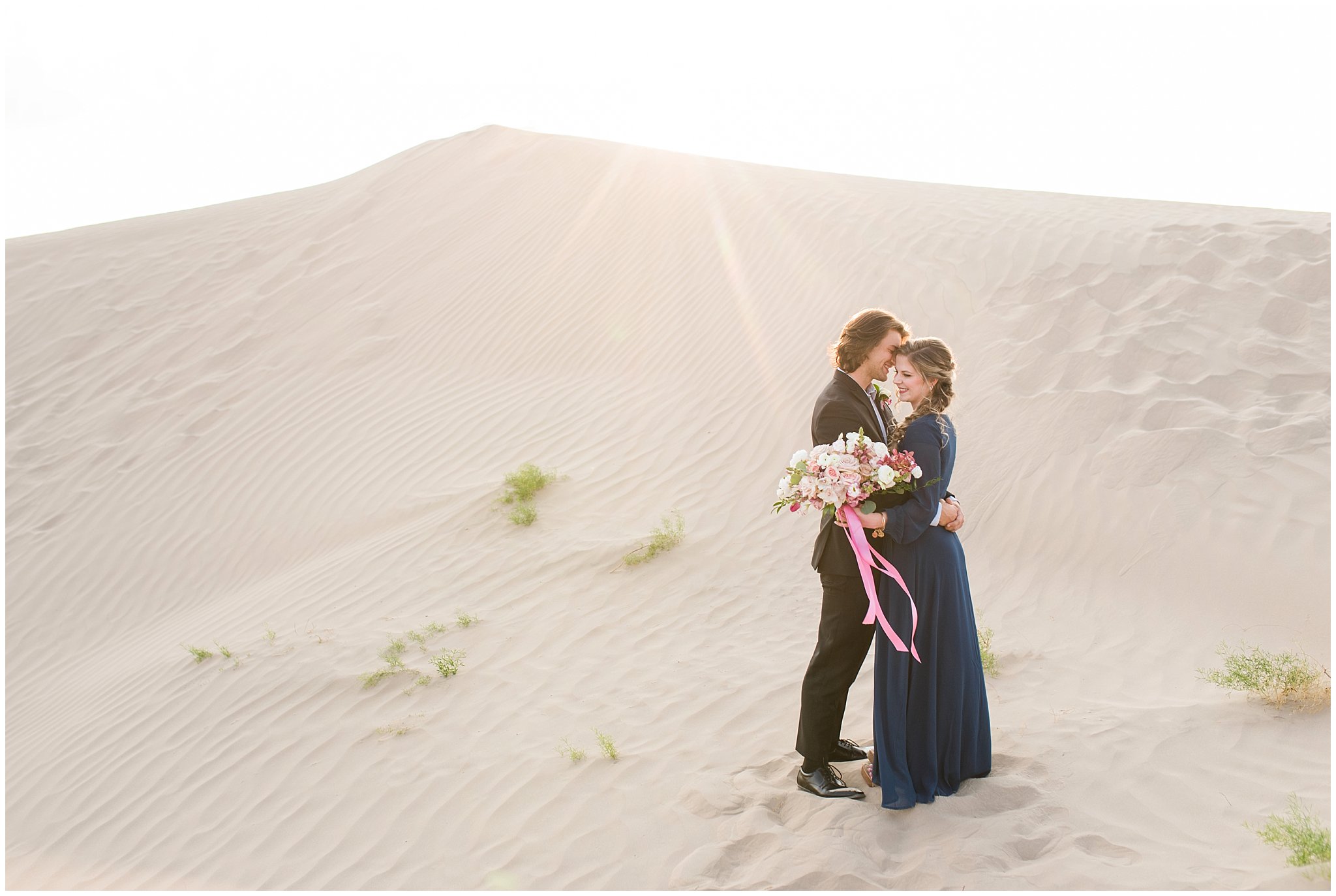 Couple dressed in black suit and blue flowy dress with white and pink orchid floral bouquet during couples session | Little Sahara Sand Dunes Milky Way Engagement Session