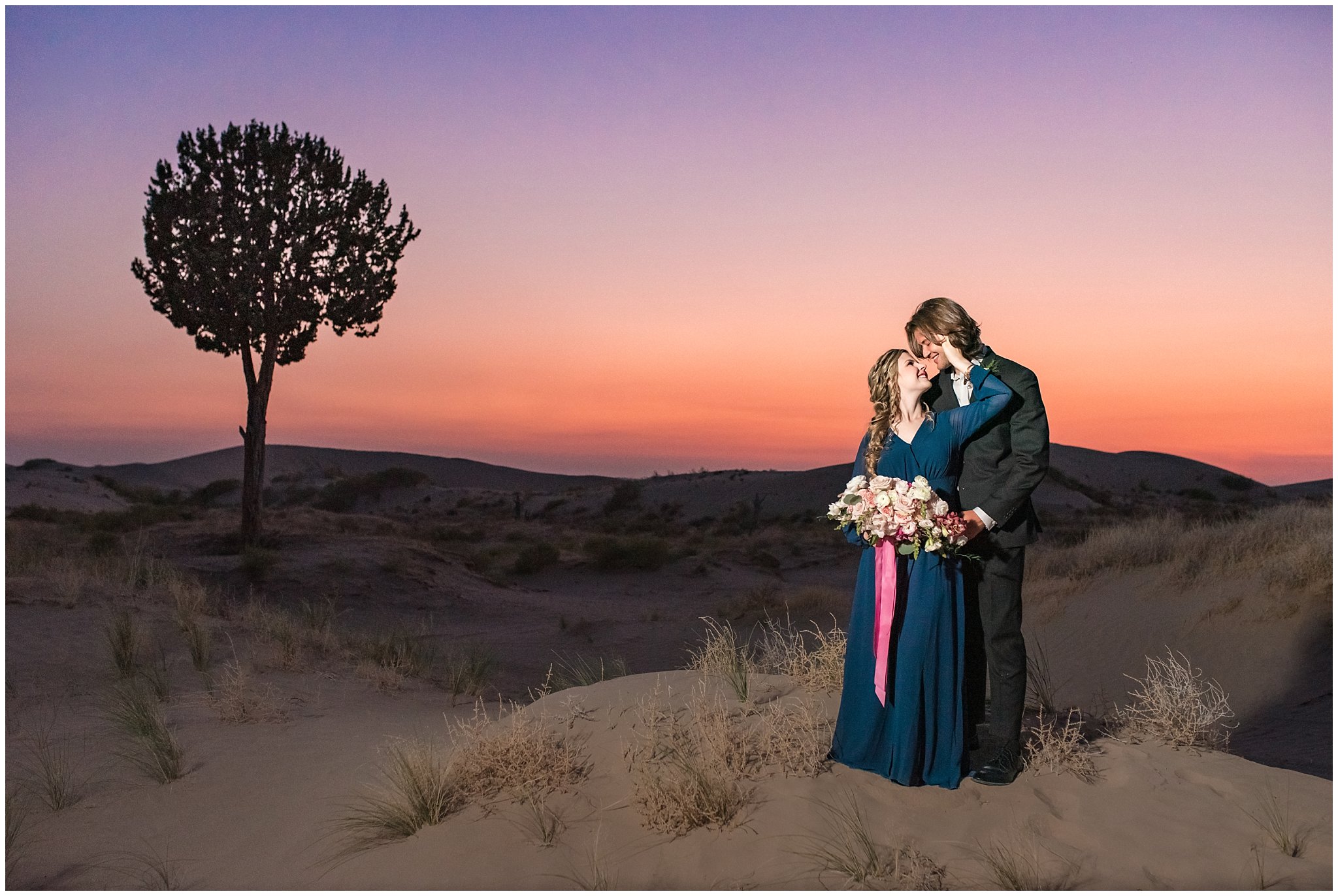 Couple at sunset dressed in black suit and blue flowy dress with white and pink orchid floral bouquet during couples session | Little Sahara Sand Dunes Milky Way Engagement Session