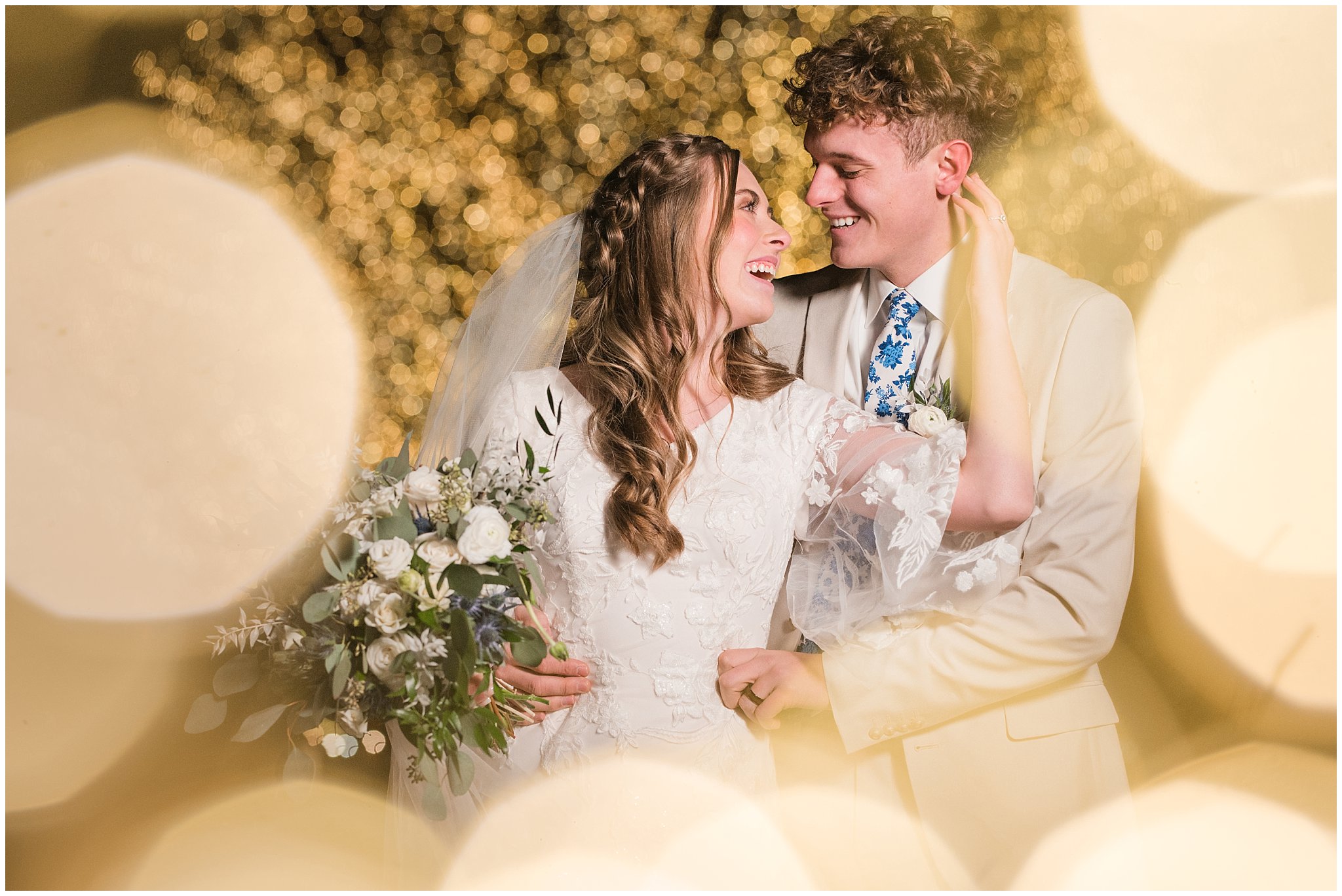 Bride and groom portraits with Christmas lights at the Tree of Life at Draper Park. Groom in cream colored suit with blue floral tie and bride with lace dress and white floral bouquet. | Oquirrh Mountain Temple and Draper Day Barn Winter Wedding | Jessie and Dallin Photography