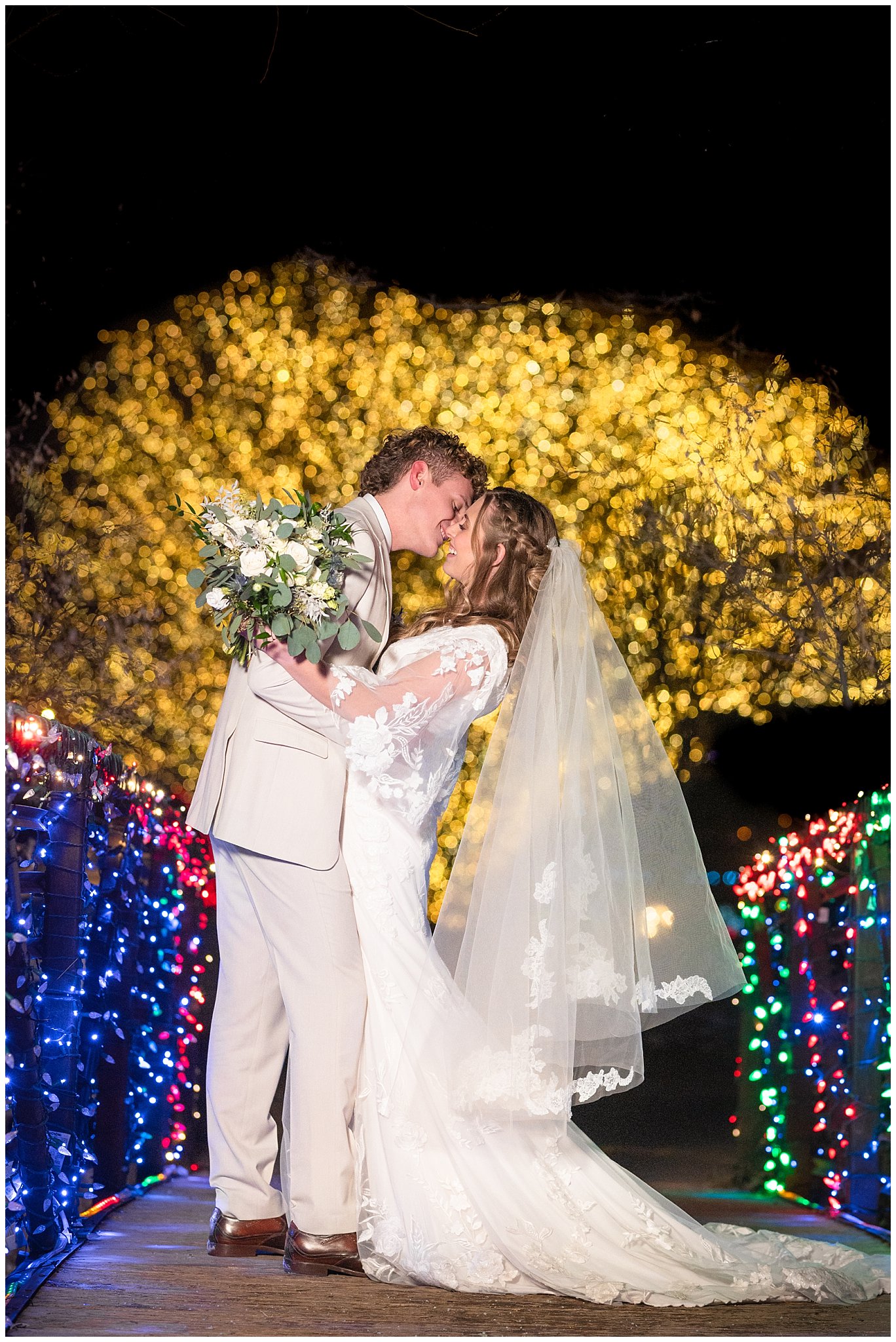 Bride and groom portraits with Christmas lights at the Tree of Life at Draper Park. Groom in cream colored suit with blue floral tie and bride with lace dress and white floral bouquet. | Oquirrh Mountain Temple and Draper Day Barn Winter Wedding | Jessie and Dallin Photography