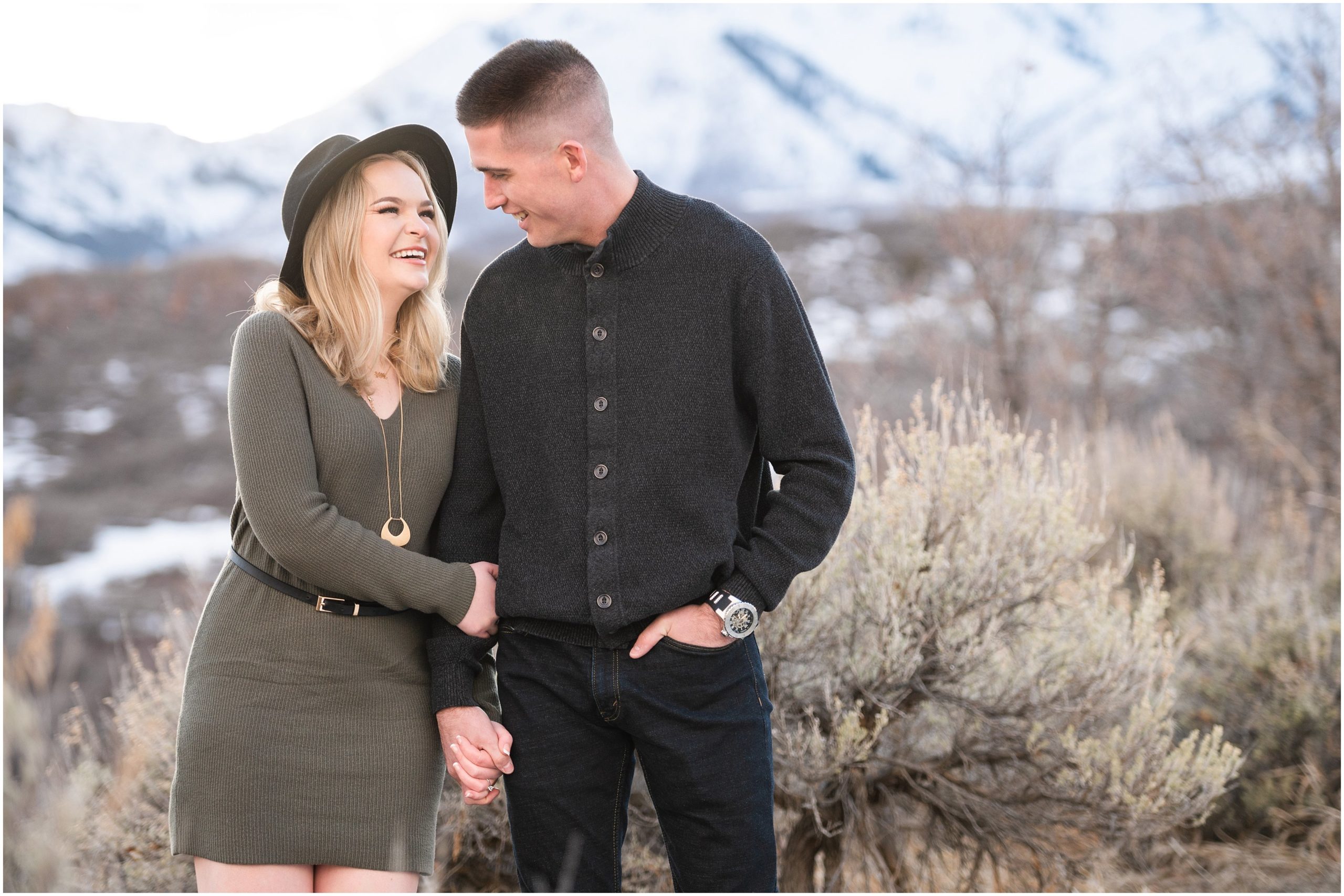 Couple in the snow during mountain destination engagement session in Utah | Wearing olive colored dress with black floppy hat and black sweater and black pants | Snowbasin Resort Snowy Engagement Session | Jessie and Dallin Photography