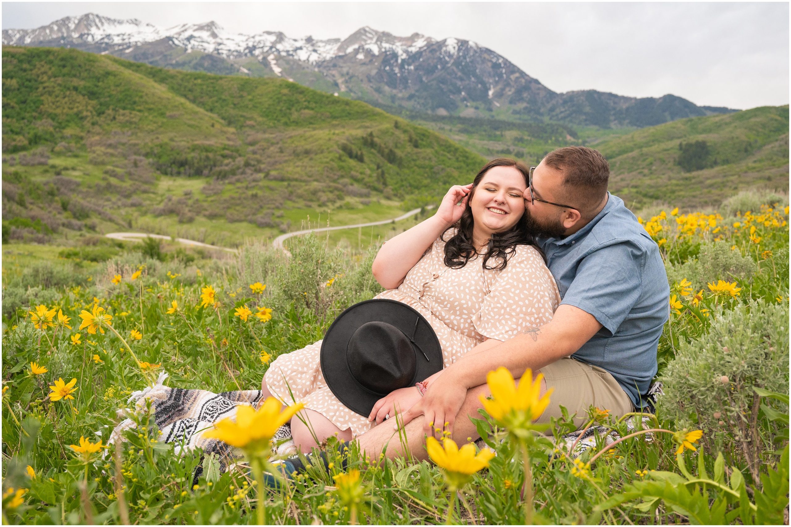 Couple during engagement session in yellow sunflower wildflowers surrounded by snowy mountain peaks | Ogden Valley Summer Engagement Session