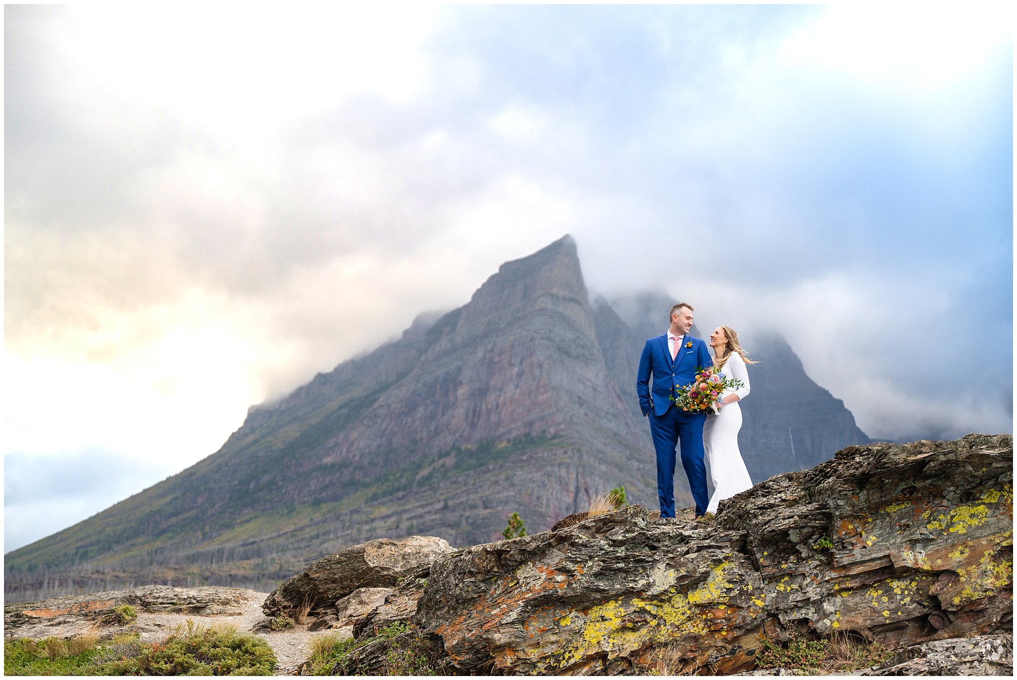 Bride and groom portraits on the Going to the Sun Road in Glacier National Park | Bride in fitted elegant dress with wildflower bouquet, and groom in blue suit with coral pink tie | Glacier National Park Wedding Formal Session | Jessie and Dallin Photography