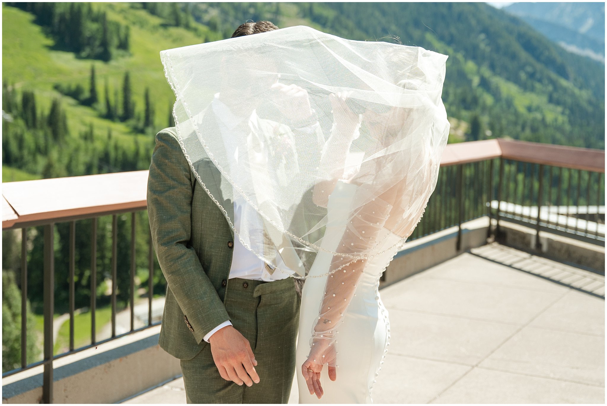 Bride and groom share moment as veil flies in front of them on the rooftop of The Cliff Lodge. Groom wearing a sage green suit and golden tie, bride in a strapless dress with gloves and veil | Snowbird Summit Summer Wedding | Jessie and Dallin Photography