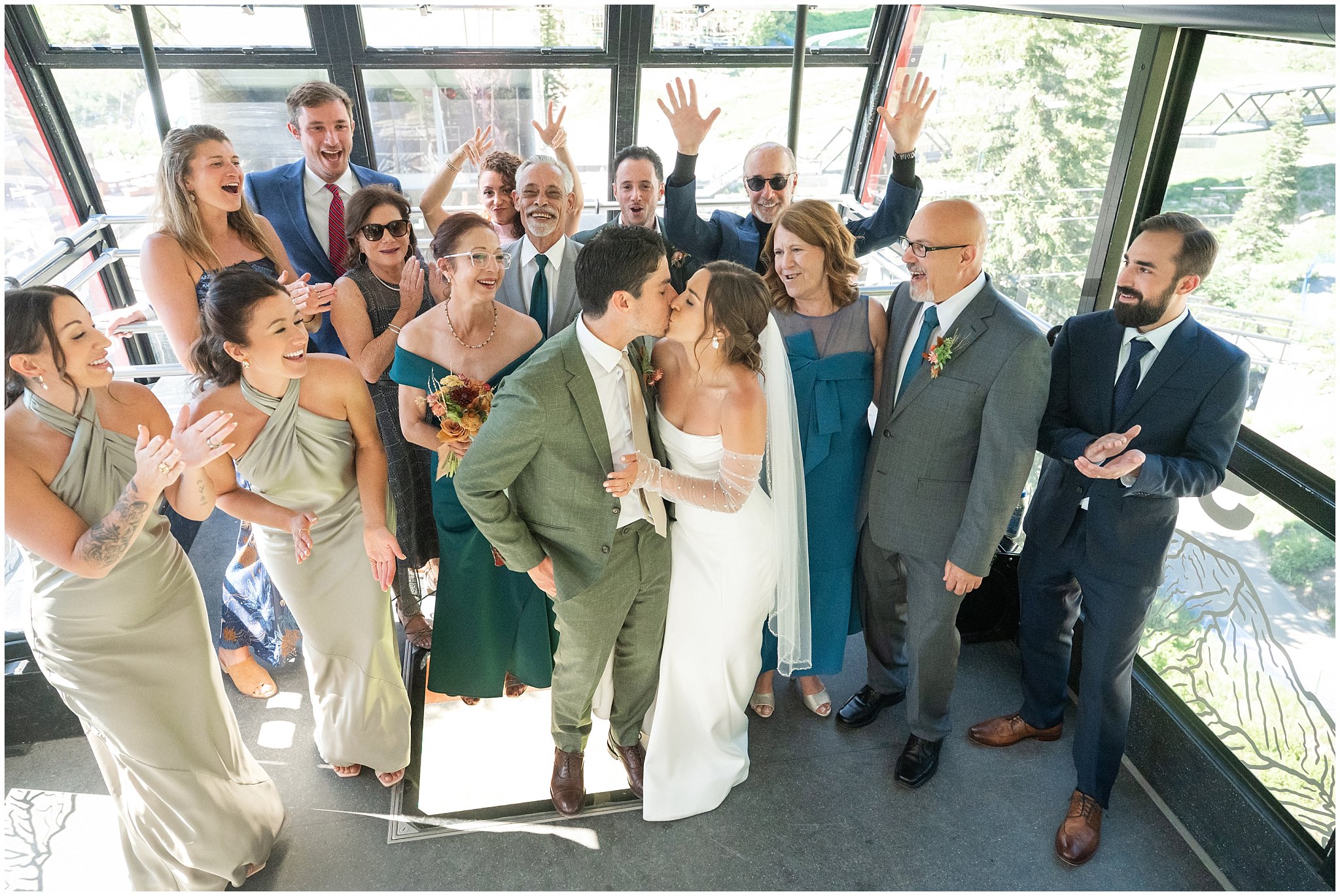 Bride and groom share a kiss surrounded by cheering guests on the Snowbird tram before their ceremony. Groom wearing a sage green suit and golden tie, bride in a strapless dress with gloves and veil | Snowbird Summit Summer Wedding | Jessie and Dallin Photography