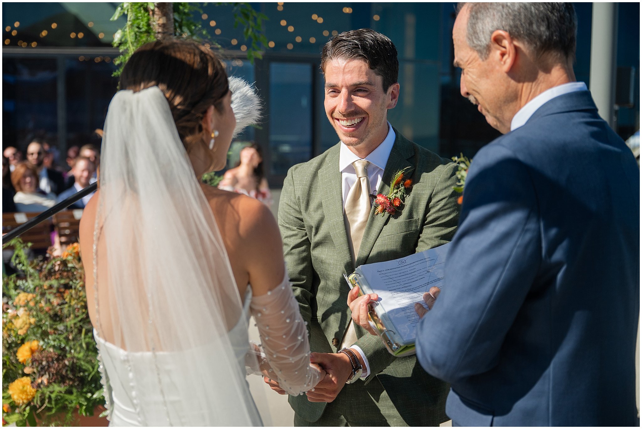 Groom laughing at the top of the aisle during ceremony. Groom wearing a sage green suit and golden tie, bride in a strapless dress with gloves and veil | Snowbird Summit Summer Wedding | Jessie and Dallin Photography