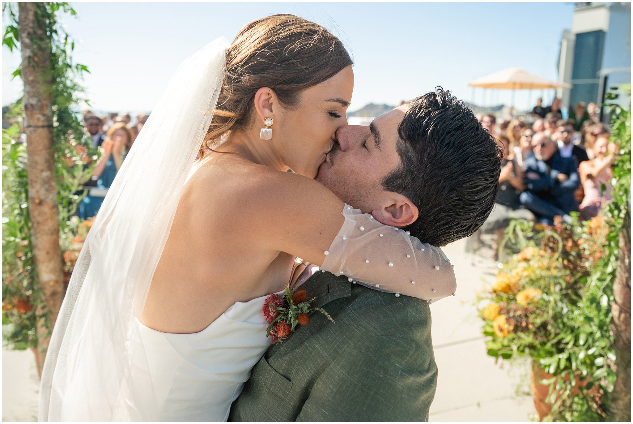 Bride and groom share their first kiss with guests in the background. Groom wearing a sage green suit and golden tie, bride in a strapless dress with gloves and veil | Snowbird Summit Summer Wedding | Jessie and Dallin Photography