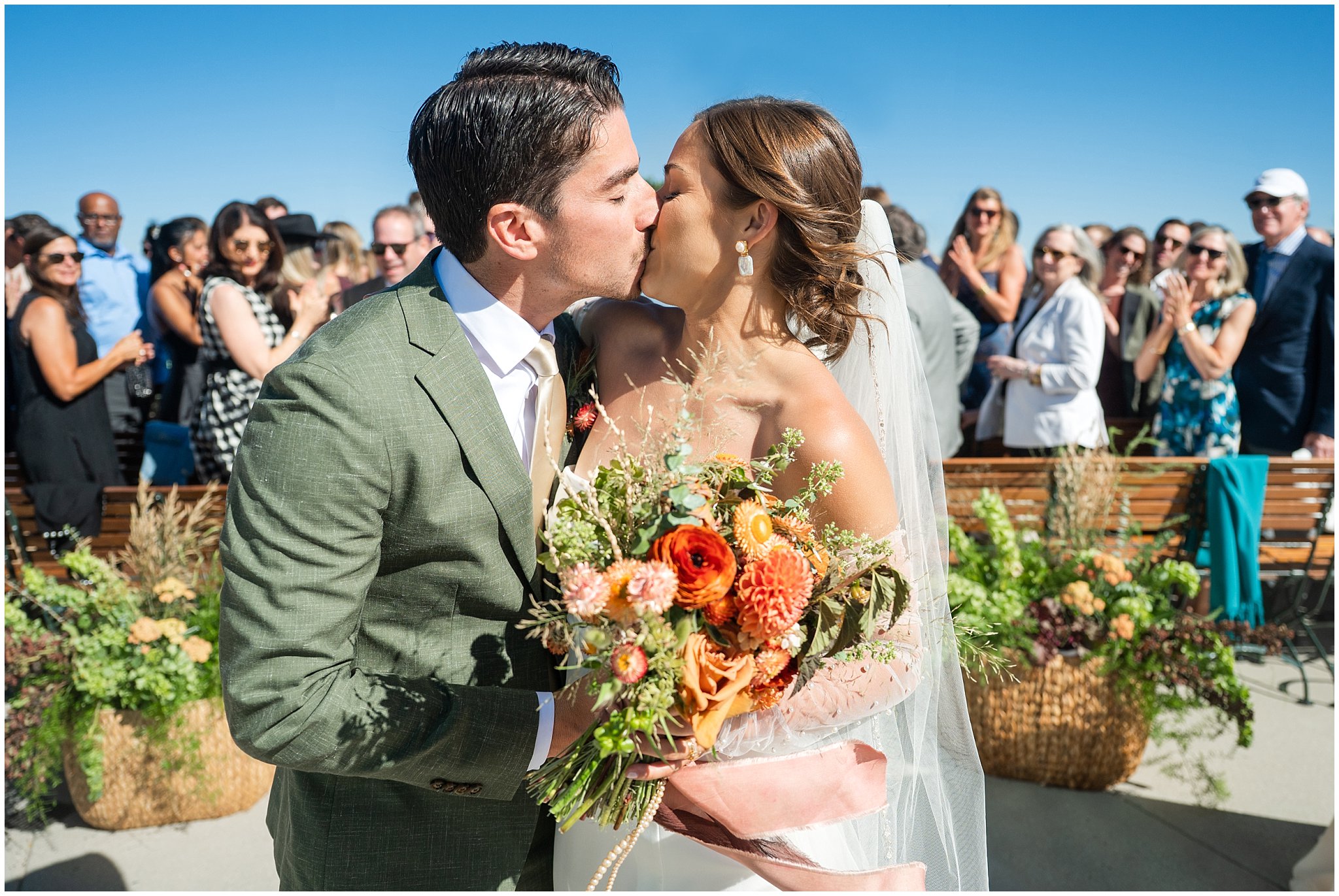 Bride and groom share their first kiss with guests in the background. Groom wearing a sage green suit and golden tie, bride in a strapless dress with gloves and veil | Snowbird Summit Summer Wedding | Jessie and Dallin Photography