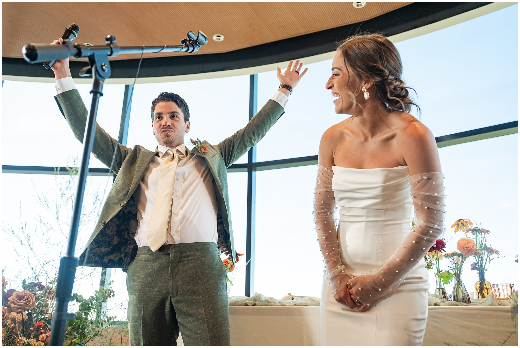 Groom pretends to drop the mic during dinner speech during their grand entrance. Groom wearing a sage green suit and golden tie, bride in a strapless dress with gloves and veil | Snowbird Summit Summer Wedding | Jessie and Dallin Photography