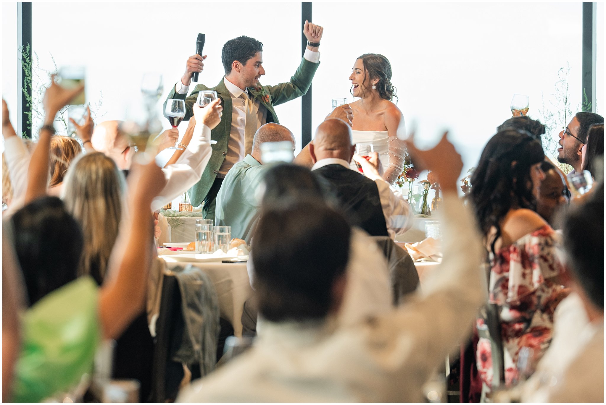 Groom raises his arms to celebrate and he and bride laugh during speech during their grand entrance. Groom wearing a sage green suit and golden tie, bride in a strapless dress with gloves and veil | Snowbird Summit Summer Wedding | Jessie and Dallin Photography