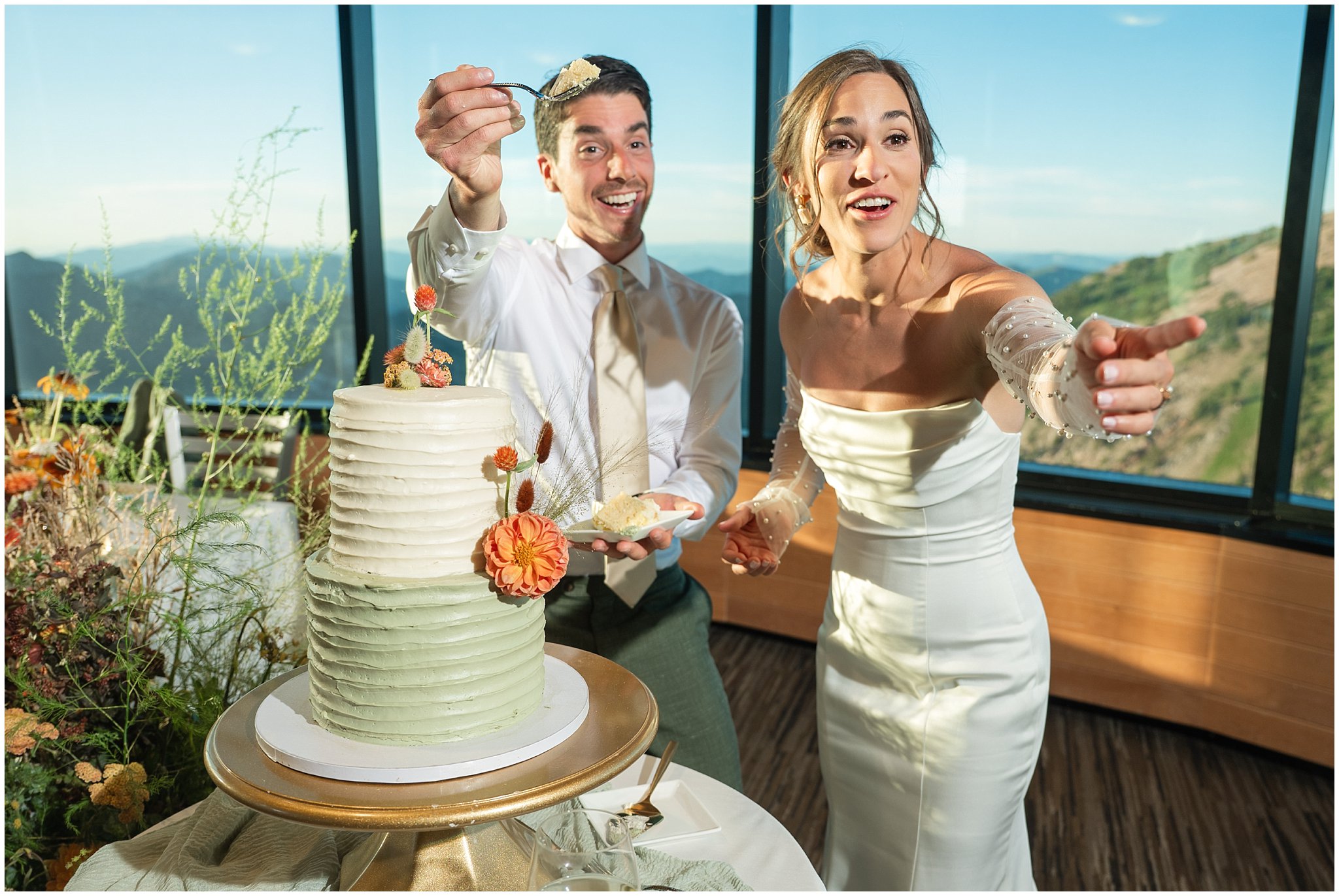 Bride and groom laugh during cake cutting overlooking the mountains. Groom wearing a sage green suit and golden tie, bride in a strapless dress with gloves and veil | Snowbird Summit Summer Wedding | Jessie and Dallin Photography