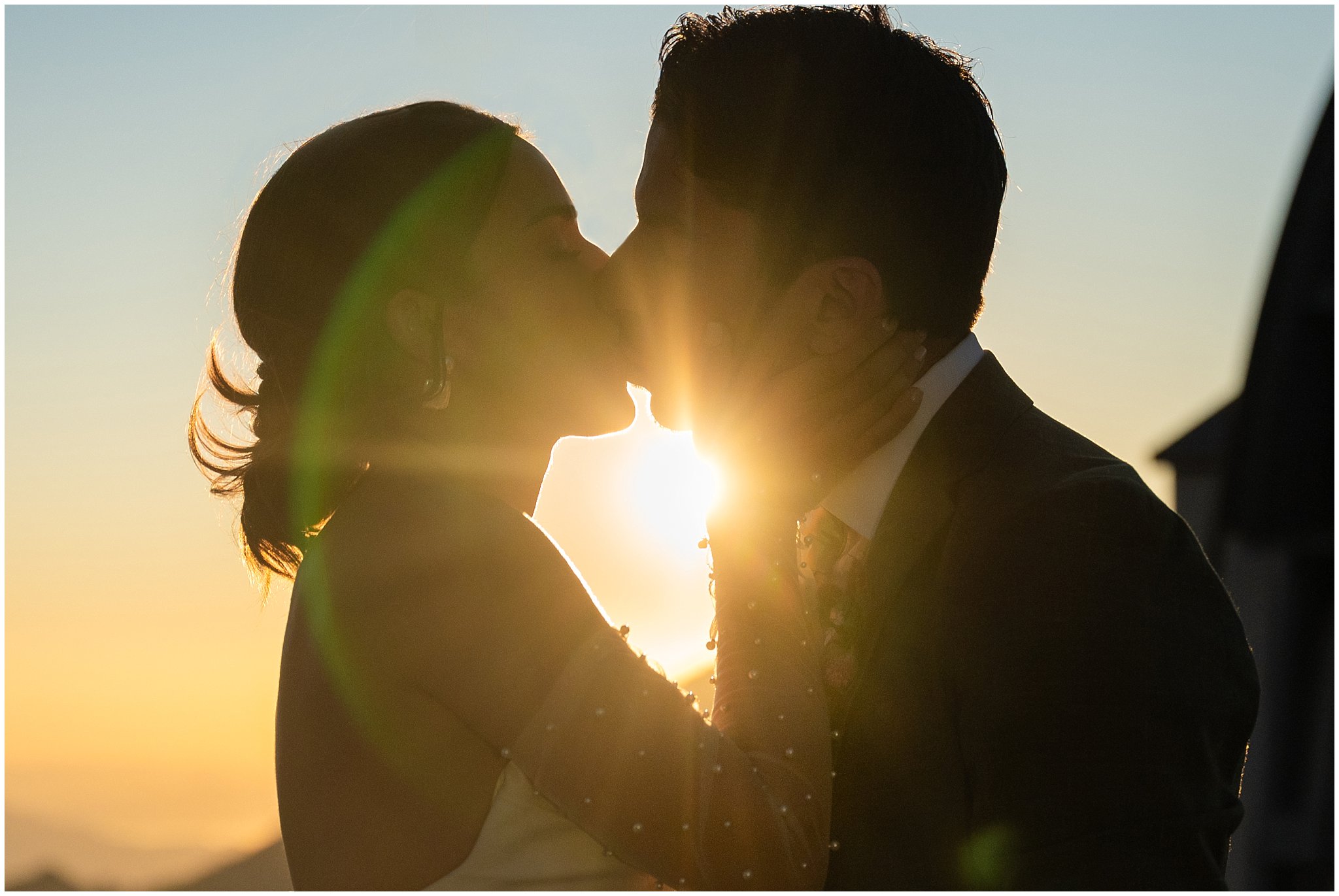 Bride and groom kissing silhouette with sun flare. Groom wearing a sage green suit and golden tie, bride in a strapless dress with gloves and veil | Snowbird Summit Summer Wedding | Jessie and Dallin Photography