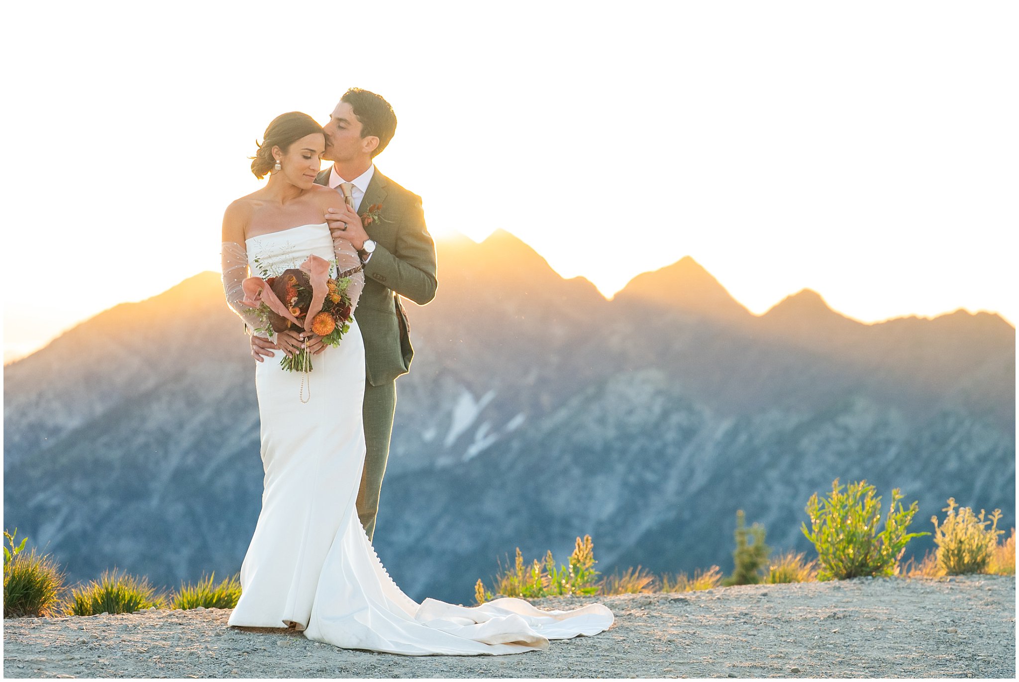 The bride and groom share moments together surrounded by the mountain peaks and a golden sunset and alpen glow. Groom wearing a sage green suit and golden tie, bride in a strapless dress with gloves and veil | Snowbird Summit Summer Wedding | Jessie and Dallin Photography